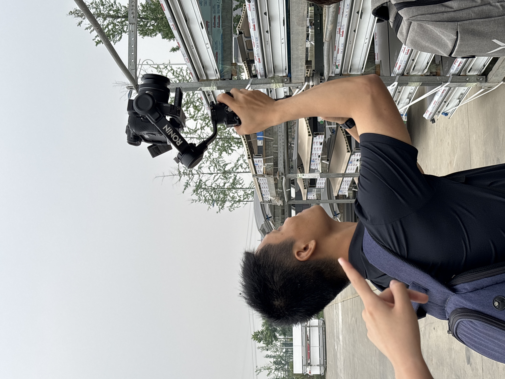 The image shows a man in Chengdu, China, holding a camera mounted on a DJI Ronin gimbal. He is filming white trim pieces stacked vertically on metal racks. The sky is overcast. The man is wearing a black shirt and a blue backpack. Another hand is pointing at the man. The location appears to be an outdoor storage area or warehouse.