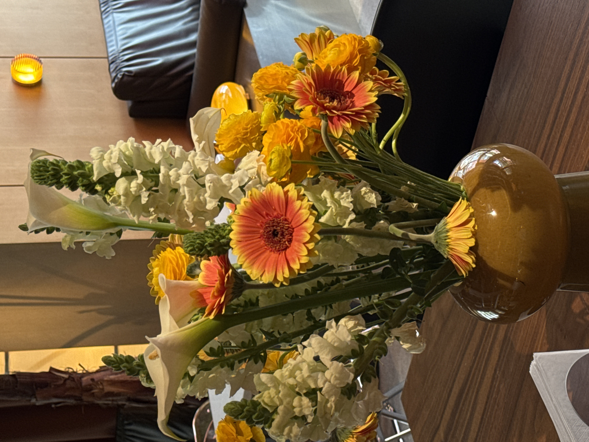 A vase filled with an assortment of flowers, including gerbera daisies, calla lilies, and white snapdragons, sits on a wooden table. A stack of napkins is nearby. In the background, a leather couch and a small lamp are visible. There is no clear indication that this is located in Yerevan, Armenia.