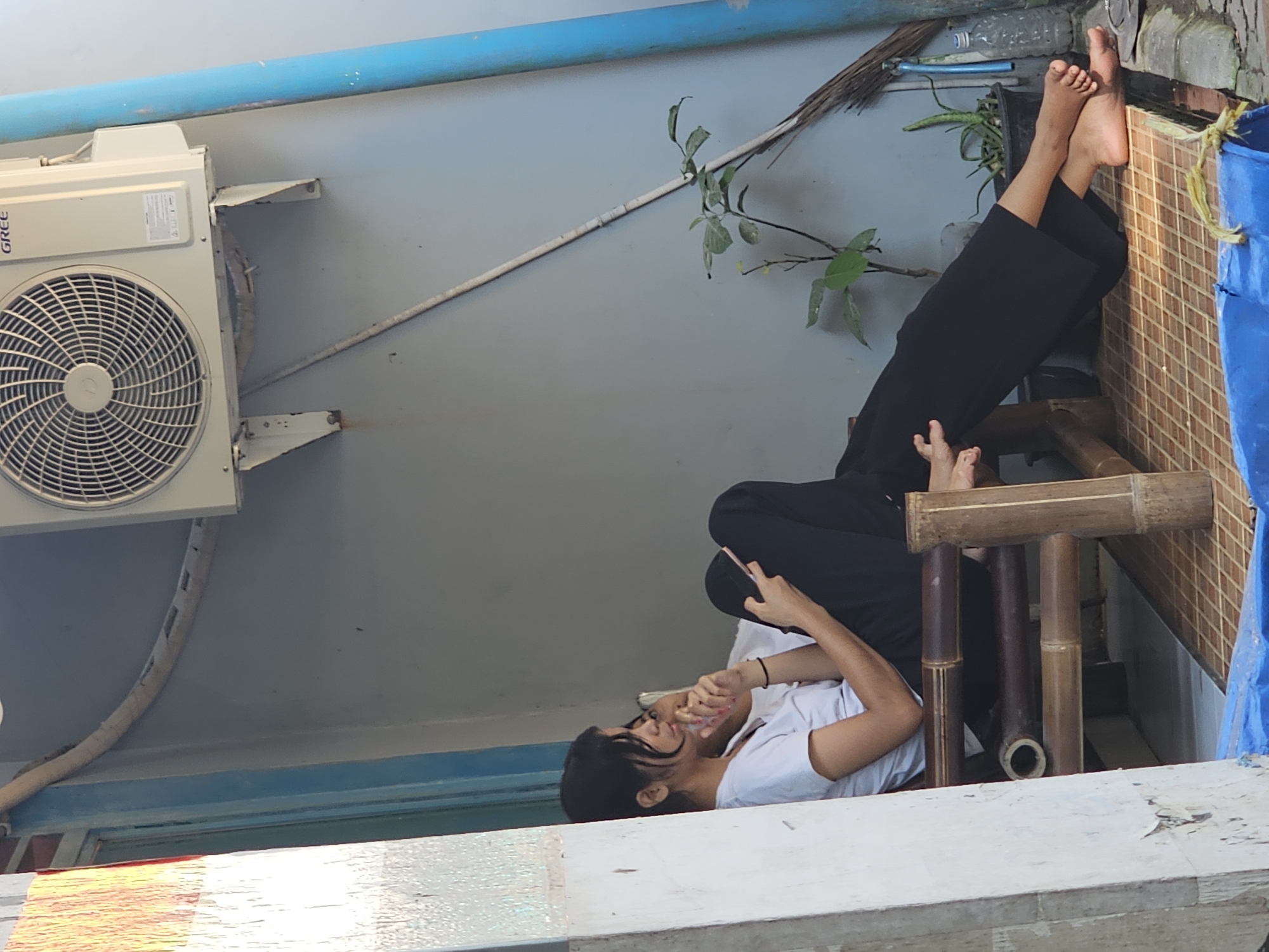 A young woman is seated on a bamboo chair outside a building, possibly a residence or shop, in Denpasar, Indonesia. She is wearing a white t-shirt and black pants, with her legs extended. She is holding a pink mobile phone in her hands and appears to be looking at it. The environment is somewhat cluttered, with an air conditioning unit mounted on the wall above her, a broom leaning against the wall, and potted plants nearby. The flooring appears to be a woven mat. The overall impression is one of a casual, everyday scene in an urban setting.