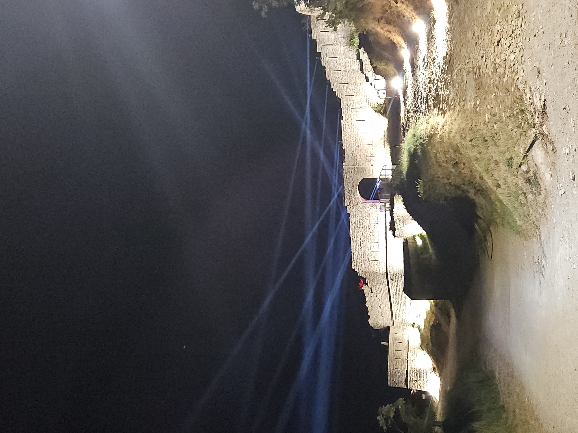 This is a nighttime, low-angle shot of a stone fortress or castle illuminated by spotlights, with several blue light beams projected into the dark sky from behind the structure. The fortress wall is made of large, rough-hewn stones and features a series of crenellations along its top. An arched entrance is visible on the left side of the fortress. The ground in front of the fortress is a dirt path, with grassy embankments on either side. Several small, bright lights are strategically placed along the base of the fortress wall and on the hillside, illuminating the structure and its surroundings. The sky above is dark and clear, with only a few faint light beams visible. There is a small, indistinct red object on top of the fortress wall near the center.