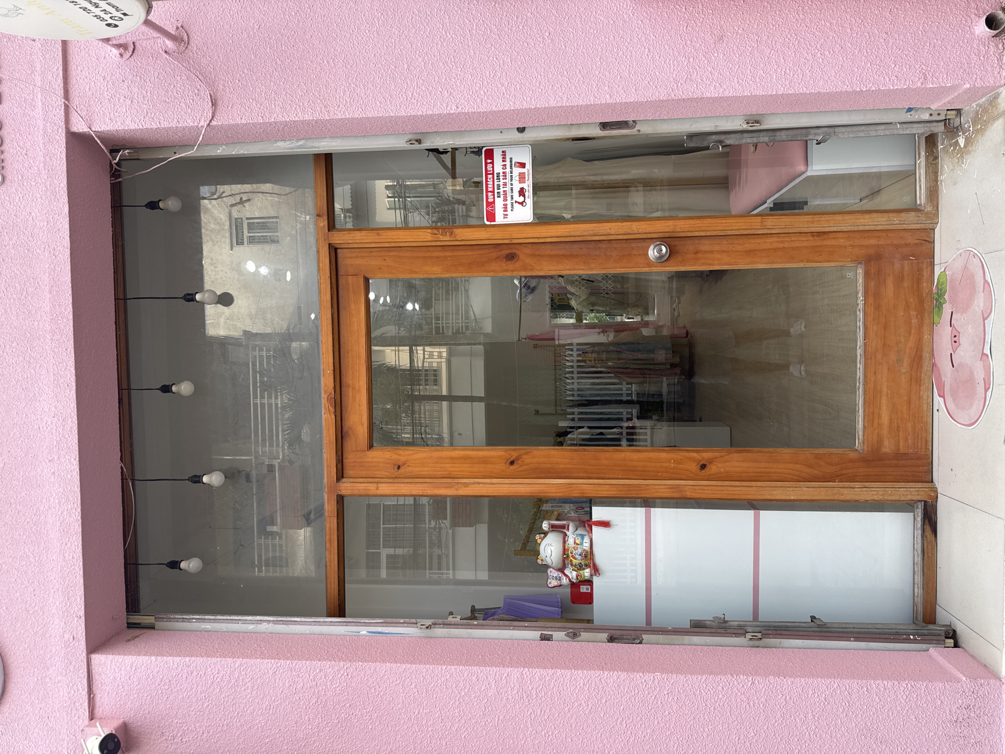 The image depicts the entrance to a pink, textured building in Nha Trang Ward, Vietnam. The entrance features a double wooden door with glass panels. To the left of the door, a large glass window reveals the interior of a shop, displaying clothing racks and a white counter with a decorative lucky cat figurine. Above the door, a red and white sign is affixed to the glass, with text in Vietnamese and English stating, "QUY KHÁCH LƯU Ý / XIN VUI LÒNG / TỰ BẢO QUẢN TÀI SẢN CÁ NHÂN / PLEASE TAKE CARE OF YOUR BELONGINGS". The exterior of the building is painted a vibrant pink, and a security camera is visible in the upper left corner. A pink pig-shaped doormat lies on the tiled ground in front of the entrance. The overall environment suggests a retail establishment.