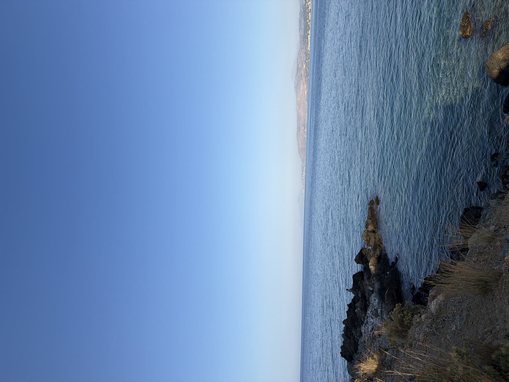 A wide shot captures a serene seascape off the coast of Georgioupoli, Greece. The vast, clear blue sky dominates the upper portion of the image, transitioning to a tranquil blue sea below. The water is calm, with subtle ripples reflecting the sunlight. Along the left side of the frame, a rocky shoreline is visible, covered in dry grasses and sparse vegetation. Several dark, rough-textured rocks protrude from the water near the shore. In the mid-ground, on the rocks, a lone figure is engaged in fishing, holding a fishing rod. In the far distance, along the horizon, a hazy coastline with hills and what appears to be a settlement is discernible. The overall atmosphere is one of peacefulness and natural beauty.