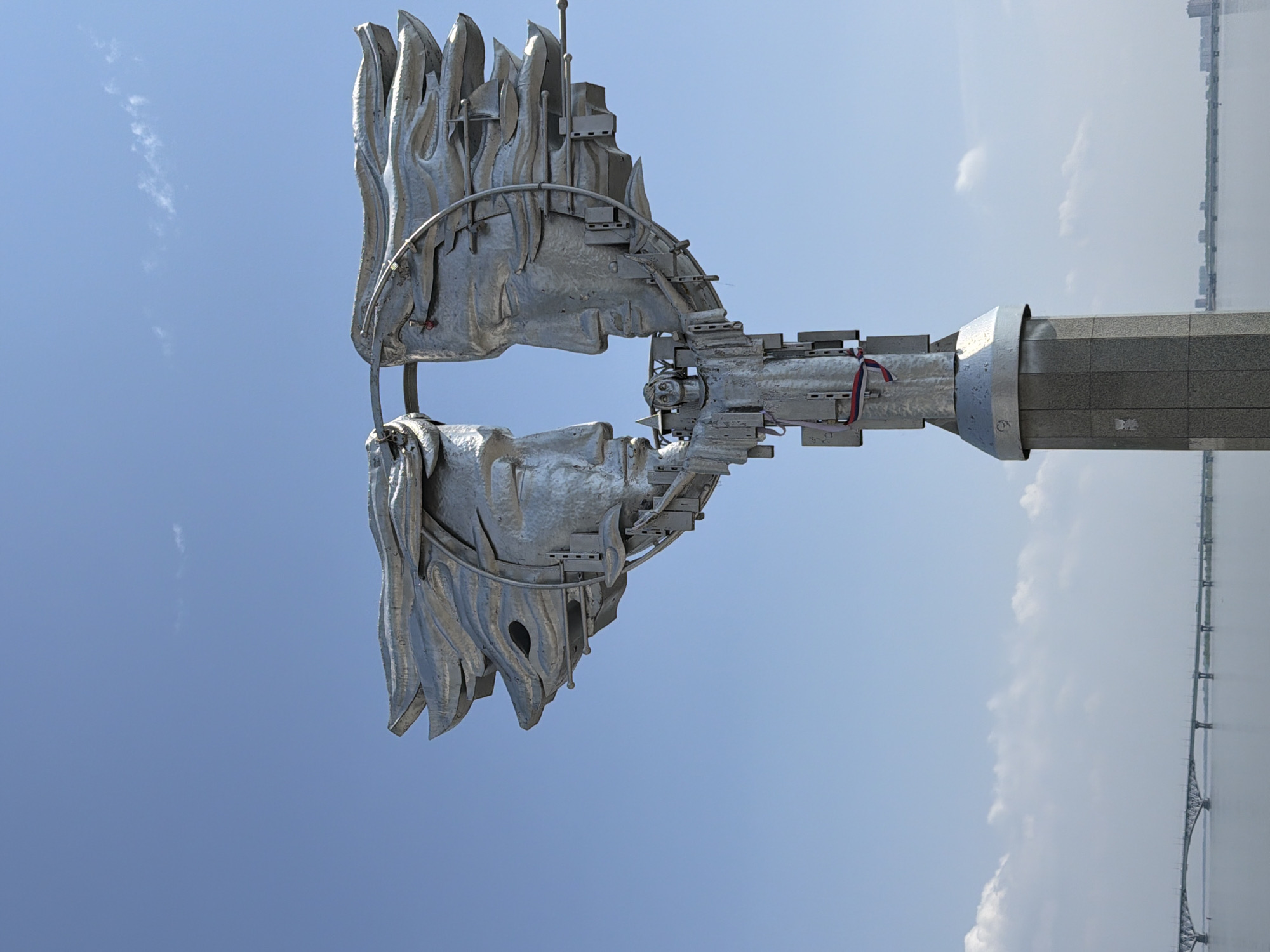 The image shows the Monument to the First Teacher in Saratov, Russia. This monument is a modern metal sculpture depicting a book or scroll with pages that resemble flames. It's mounted on a tall, rectangular column with a light gray, segmented texture. The sky is light blue with a few scattered clouds. In the distance, a large body of water is visible, with a long bridge extending across it. The image appears to have been taken from a low angle, looking up at the monument.