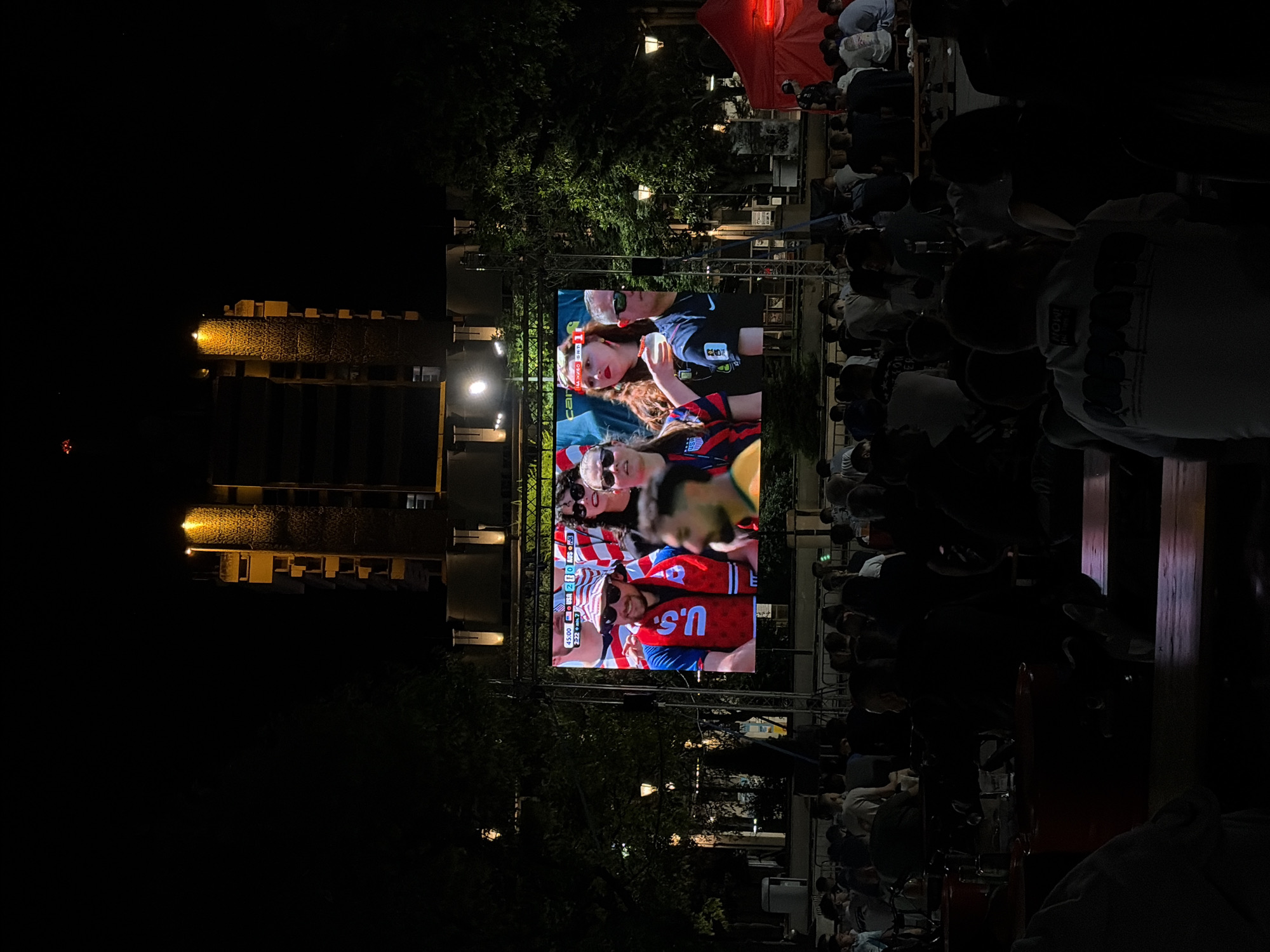 An outdoor night scene depicts a large digital screen displaying a soccer match, likely the USA vs. Australia game based on the on-screen graphics and the attire of the people on screen. A crowd of spectators is visible in the foreground, seated at tables and standing, facing the screen. The background features a building with illuminated windows and trees. A red tent is set up to the right of the screen. The lighting suggests it's evening or night, with the screen and building lights being the primary light sources. The overall atmosphere appears to be that of a public viewing event.