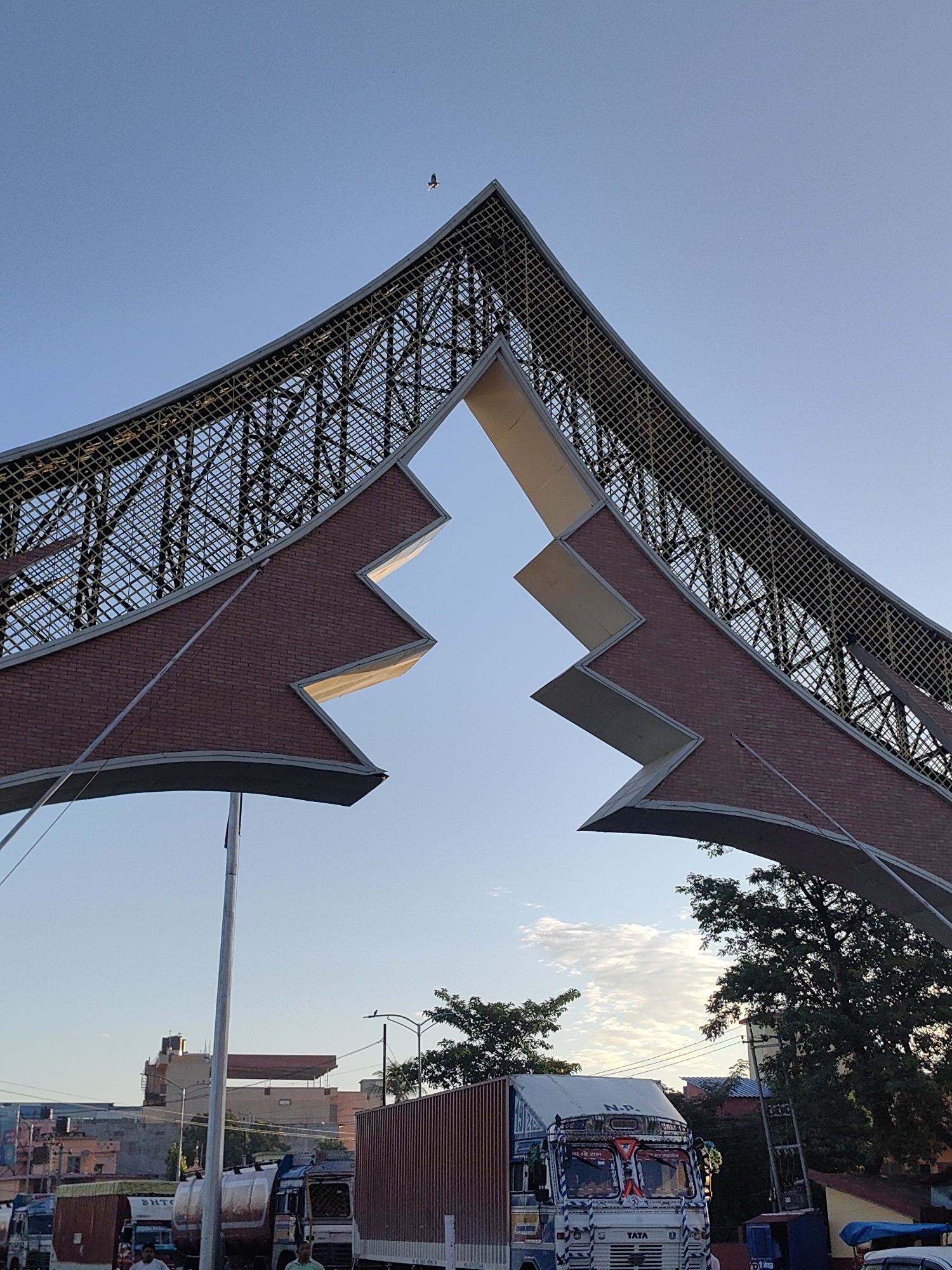 The image captures the Belahiya border crossing in Nepal, characterized by a distinctive architectural archway. The arch is constructed with a steel lattice structure supporting a stepped brick facade. The sky above is clear blue, with a few wispy clouds. A bird is visible in flight high in the sky.
Below the arch, a busy street scene unfolds. Several large trucks are parked or moving, including a white Tata truck adorned with colorful decorations and Hindi text, and a large container truck. A silver tanker truck is also present. People are visible near the vehicles, though their features are not distinct. Utility poles and streetlights dot the background, along with various buildings, suggesting a commercial or urban environment. Green trees are interspersed throughout the scene, with one large tree prominent on the right side. The overall impression is of a functional border point with notable architectural elements.