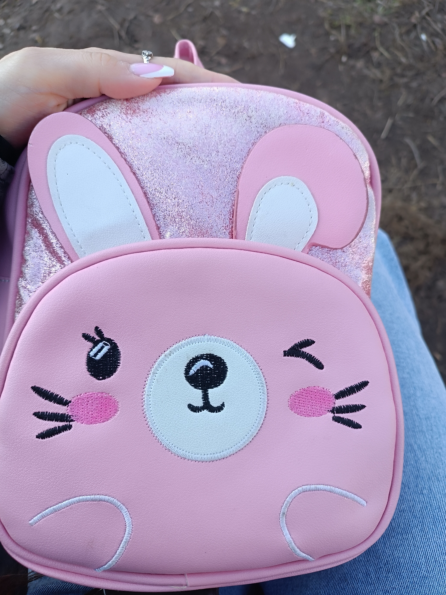 A close-up shot features a pink backpack with a bunny face design, held in a person's hand. The backpack has bunny ears, one with a white inner section. The bunny face includes a circular nose with stitched details, whiskers, and eyes, one of which is winking. The bag is resting on a pair of denim jeans. A hand is visible, with a ring on the thumb. The background is out of focus, showing patches of ground. The location and city are unknown.
