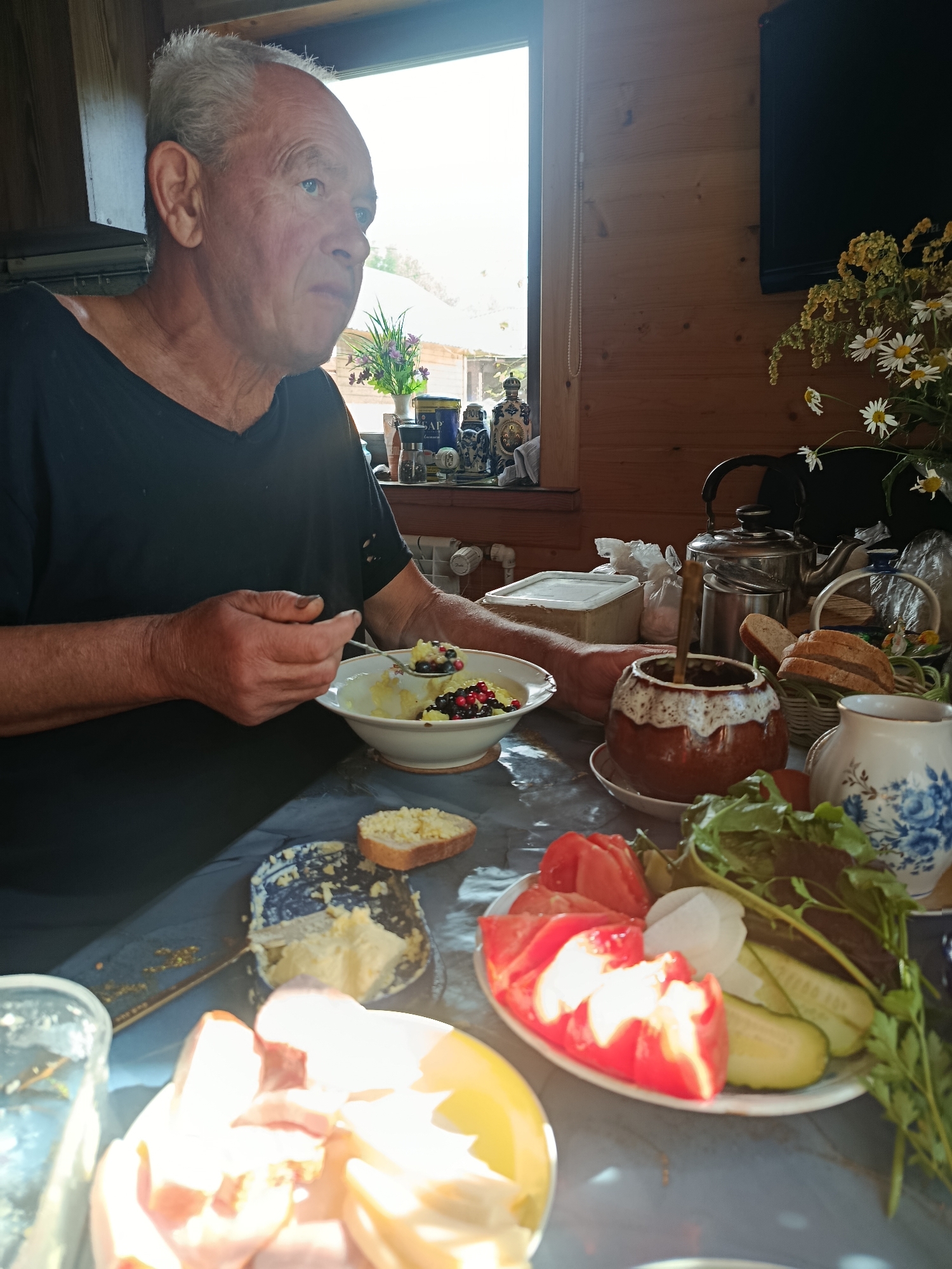 An elderly Caucasian man with grey hair is seated at a table, eating from a bowl. He is wearing a black t-shirt and has a spoon in his right hand, lifting food to his mouth. The table is set with various food items, including a plate of sliced tomatoes, cucumbers, and greens, a bowl of what appears to be porridge with berries, bread, and a dish of butter. A rustic ceramic pot and a metal teapot are also visible on the table. The background suggests an indoor setting with wooden walls and a window showing a bright, sunlit exterior. The overall scene depicts a simple meal in a possibly rural or rustic environment.