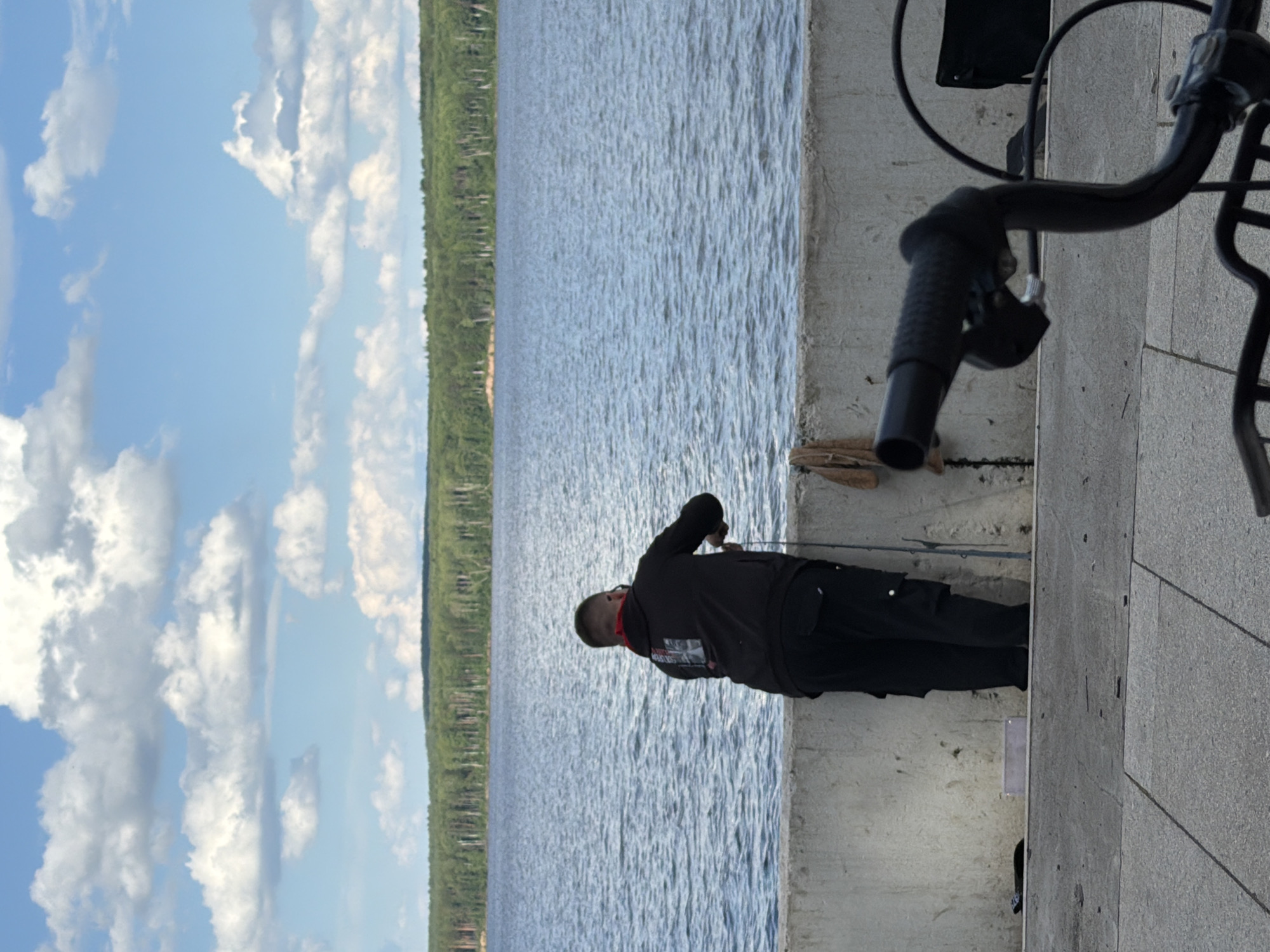 The image captures a scene at the embankment of the Kama River in Chaikovsky, Russia. A solitary individual, seen from behind, is fishing from a concrete barrier. The person is wearing a dark hooded jacket and black cargo pants, with a red bandana or scarf visible around their neck. A fishing rod is held in their hands, extending towards the water. The river stretches out in front, with a dense forest bordering the opposite bank. The sky above is filled with scattered white clouds against a bright blue backdrop. In the foreground, the handlebars and part of the frame of a bicycle are visible, suggesting its presence in the immediate vicinity. The ground is paved with light-colored square tiles. The overall atmosphere is tranquil and suggests a leisure activity by the water.