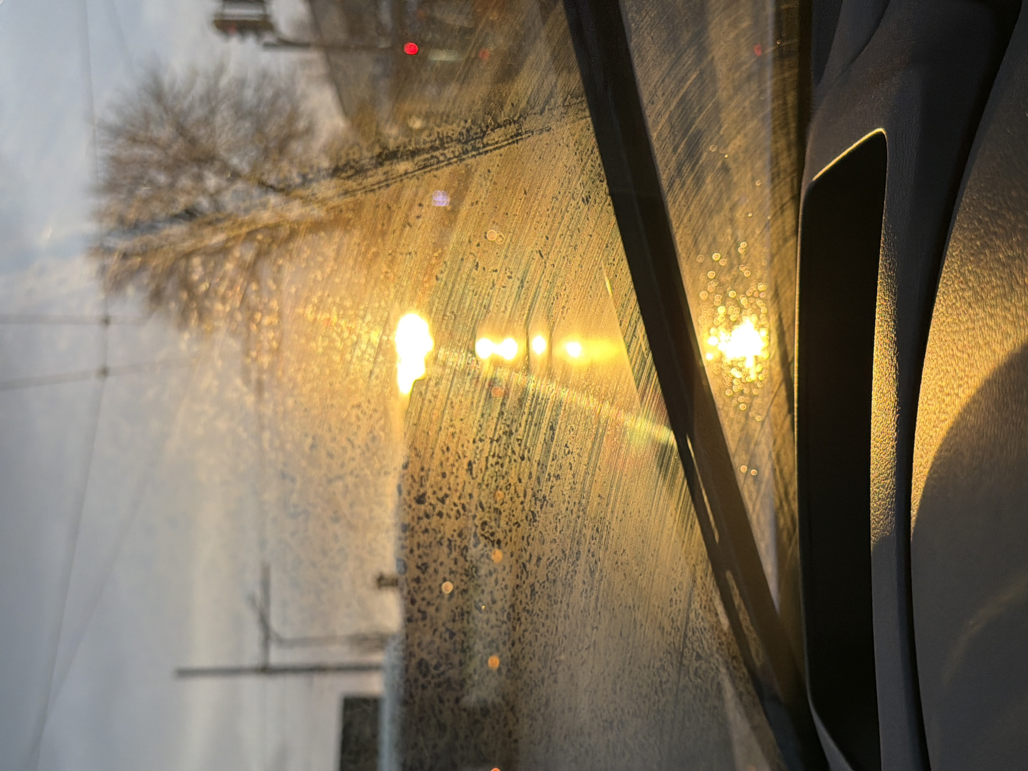 The image depicts a view through a dirty car window, likely taken in Sofia, Bulgaria. The window is streaked with grime and water spots, obscuring the outside scene. The bright sunlight shines through the window, creating glare and illuminating the interior of the car. Outside, a blurred tree and streetlights are visible. The interior of the car is visible in the right side of the frame, including the car's interior trim and details of the door.