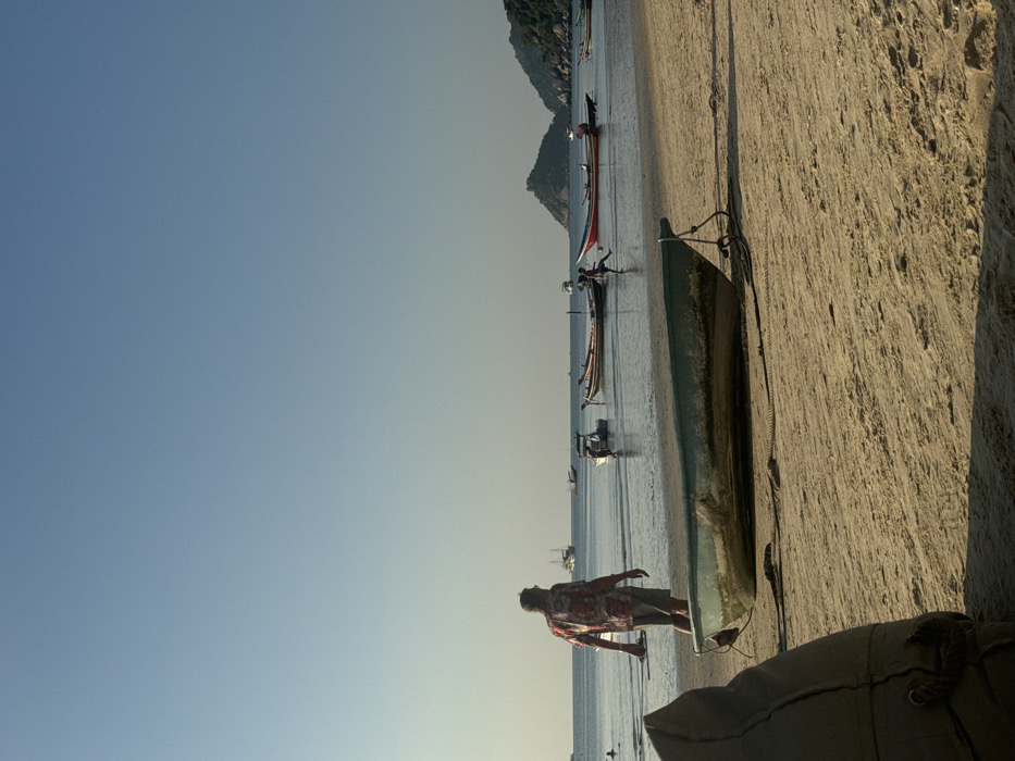 This image depicts a coastal scene under a clear, light blue sky. The location is Unknown, Unknown.
In the foreground, the lower left quadrant contains a large, indistinct, light brown or beige object, possibly a beanbag chair or cushion, partially visible. The immediate foreground consists of light brown sand, showing some texture and shadows. A small, weathered, teal or pale green wooden boat is beached on the sand in the mid-left, facing slightly right, secured by a rope visible in front of its bow.
In the midground, a male individual, observed from behind, walks towards the water on the sandy beach. He wears a short-sleeved, patterned shirt (red, white, and dark colors) and light-colored shorts. His shadow is cast to his left. To the right of this individual, in the shallow water, two young individuals appear to be interacting, possibly playing, with one holding a red object resembling a ball. Several longtail boats and smaller motorboats are anchored in the calm, reflective water.
The background shows a wide expanse of calm, slightly hazy ocean. On the far right, a verdant, rocky landmass or island with a prominent peak is visible. Further left, a smaller, lower island or landmass is also present. Several other boats, including larger vessels with masts, are scattered across the water towards the horizon. The sun appears to be low, casting long shadows and illuminating the scene from the right, indicating either early morning or late afternoon.