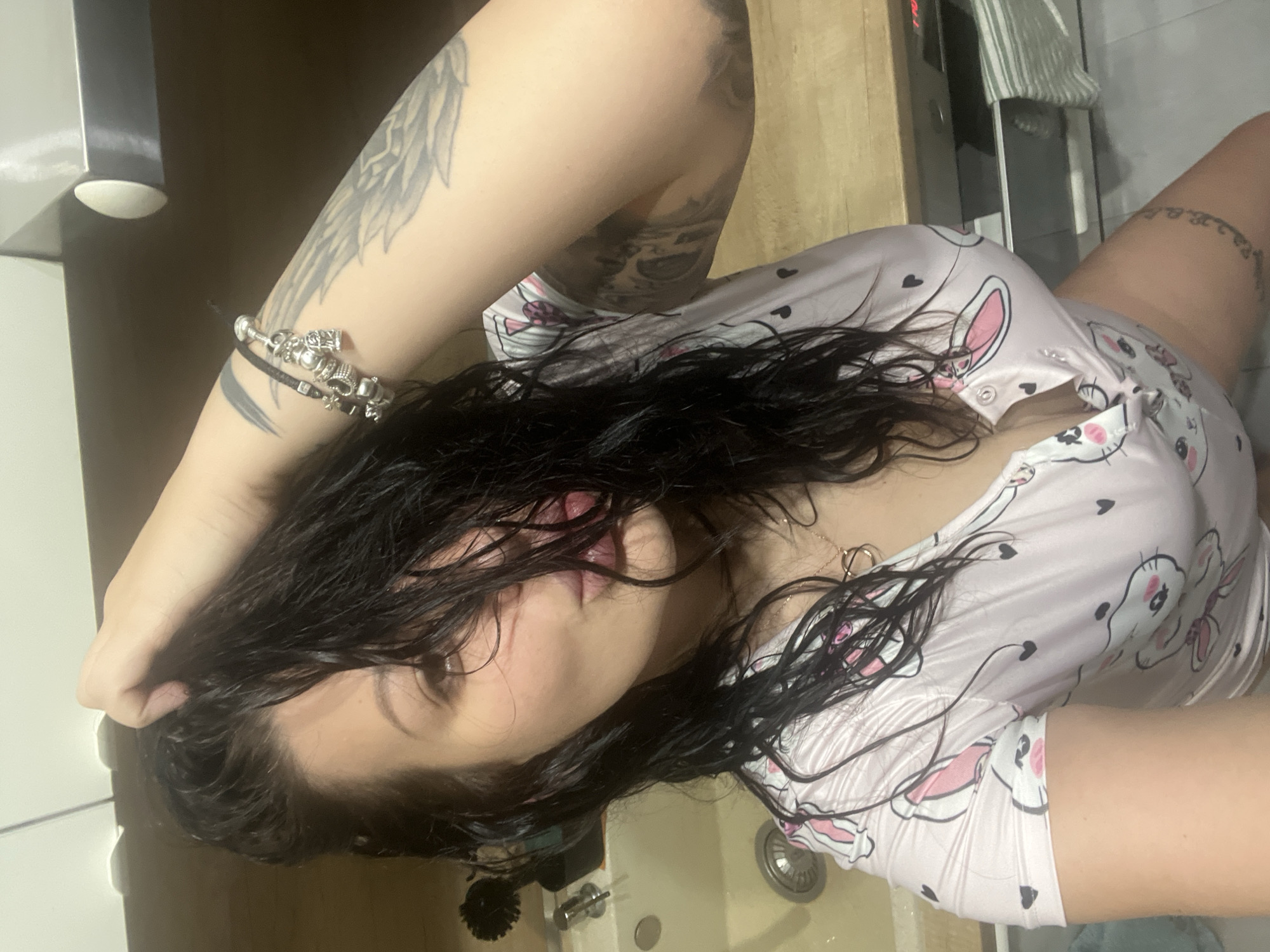 A woman with wet, dark hair is lying down, wearing a pink and white patterned top. Her left arm is raised with a bracelet visible. Tattoos adorn both arms and part of her right leg. A silver sink and faucet are visible near her head. The location is unknown, and there is no indication this photo was taken in Plovdiv, Bulgaria.