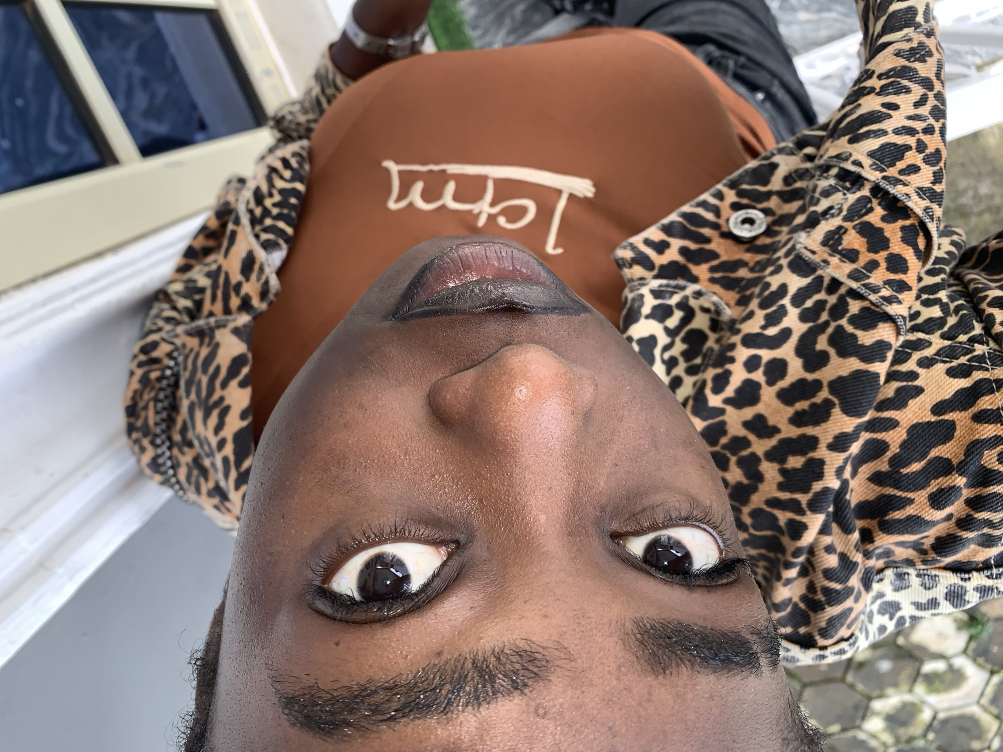 A close-up, rotated portrait of a Black individual's face and upper body is captured outdoors. The person is wearing a brown t-shirt with a cream-colored graphic and a leopard-print jacket. Their eyes are dark and alert, and their lips are painted with dark lipstick. The background suggests an outdoor setting with hexagonal paving stones visible at the bottom and a light-colored wall and windows to the upper right. The scene appears to be set in Gura Top, Nigeria.