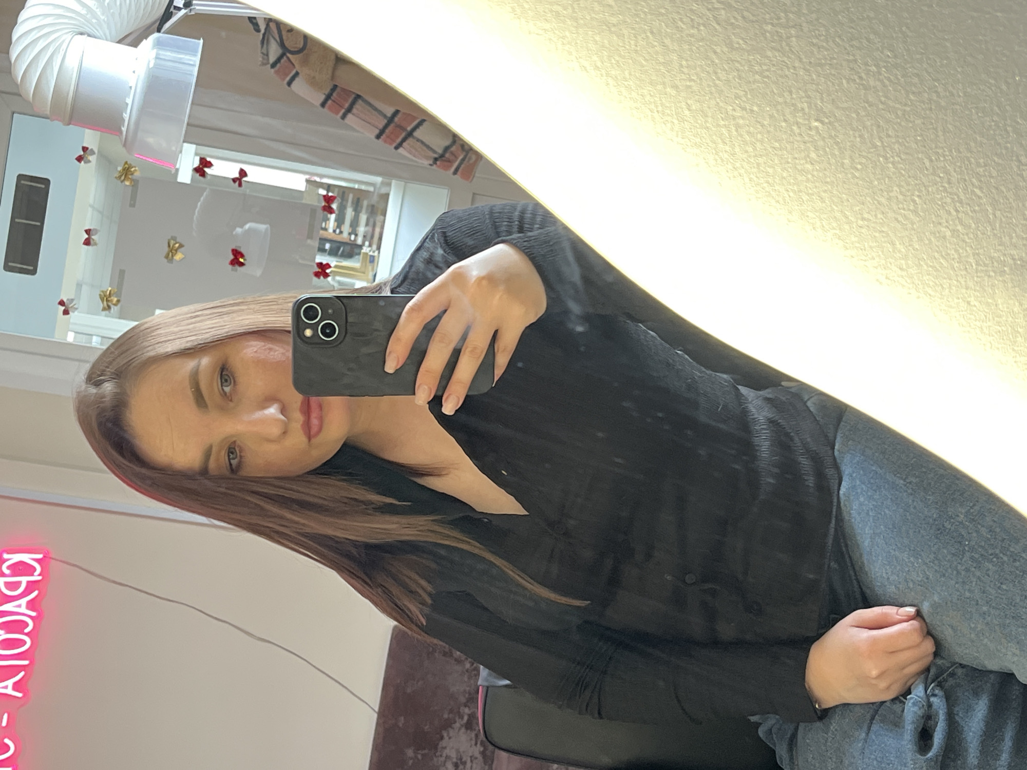 A woman is taking a selfie in a mirror. She is holding a smartphone with a dark, textured case. The woman has light brown hair and is wearing a black shirt and denim jeans. Her left hand is visible, holding the phone, and her right hand rests on her leg.
In the background, the reflection shows an interior space with a neon sign that reads “- КРАСОТА -”. There is also a ventilation system visible at the top, along with small bows stuck to the mirror and possibly the wall behind it. The location may be a salon or beauty parlor, potentially in Chaikovsky, Russia, based on the Cyrillic text of the neon sign.