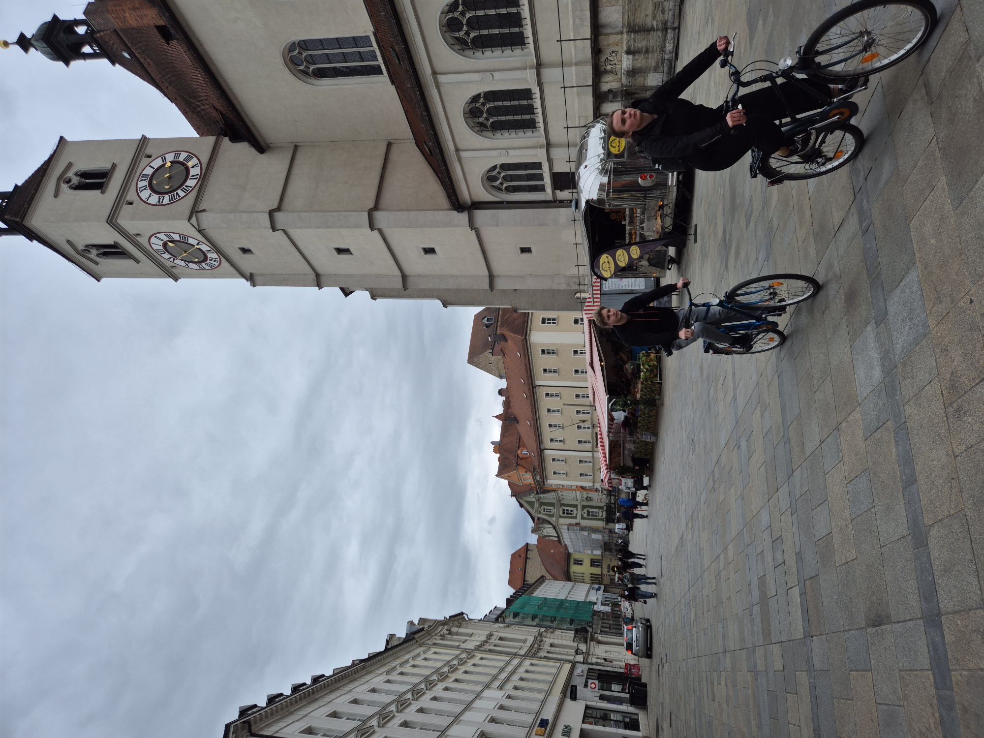 The image depicts a street scene in Regensburg, Germany, with a prominent church tower. Two young people are cycling towards the viewer on a cobblestone street lined with buildings and market stalls. The church tower, made of light-colored stone, features two clock faces with Roman numerals. The sky is overcast. In the background, people are walking, and there are several market stalls with awnings. A silver car is parked on the left side of the street. The overall impression is that of a somewhat gray, possibly cool day in an urban European setting.