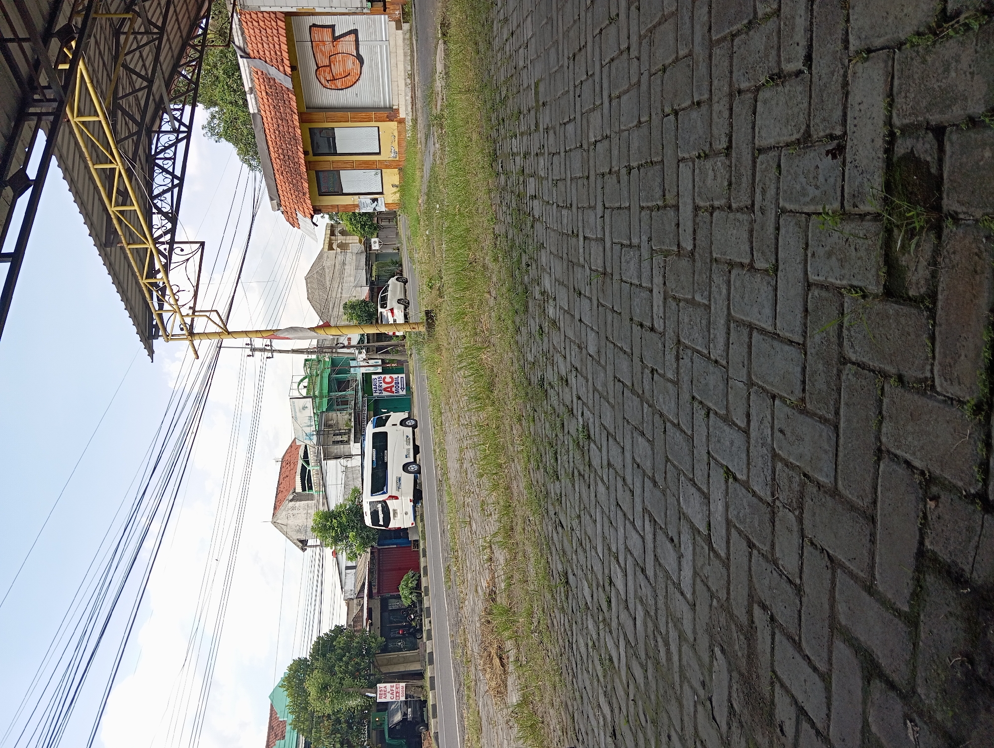 The image depicts an urban street scene in Pelem, Indonesia, under an overcast sky. In the foreground, a dark grey paved area composed of rectangular blocks extends from the bottom of the frame, with sparse green grass and weeds growing between the pavers and in an adjacent unpaved strip. A metal canopy structure with yellow support beams and a dark grey corrugated roof extends from the upper right, partially obscuring the sky.

The midground features an asphalt road with a raised median strip separating traffic lanes. A white minibus, possibly a Toyota Hiace or similar, with blue accents and visible text "Anugerah Rental," is traveling left on the road. Behind it, a white sedan is also visible, heading in the same direction. On the left side of the road, a green pickup truck is parked near a building with a visible sign for a "REST AREA CAFE." On the right side of the road, a single-story building with a red-tiled roof and yellow-painted facade features two large window frames and a closed garage-style door with orange graffiti reading "GST." An "AC Mobil" service sign is affixed to the right side of this building. A yellow utility pole, draped with an Indonesian flag, stands near the center of the frame. Numerous overhead utility lines crisscross the sky above the street.

The background shows various residential and commercial buildings with different roof styles, interspersed with trees. No individuals are visibly present or engaged in specific actions. The overall environment suggests a moderately busy commercial or residential thoroughfare.
