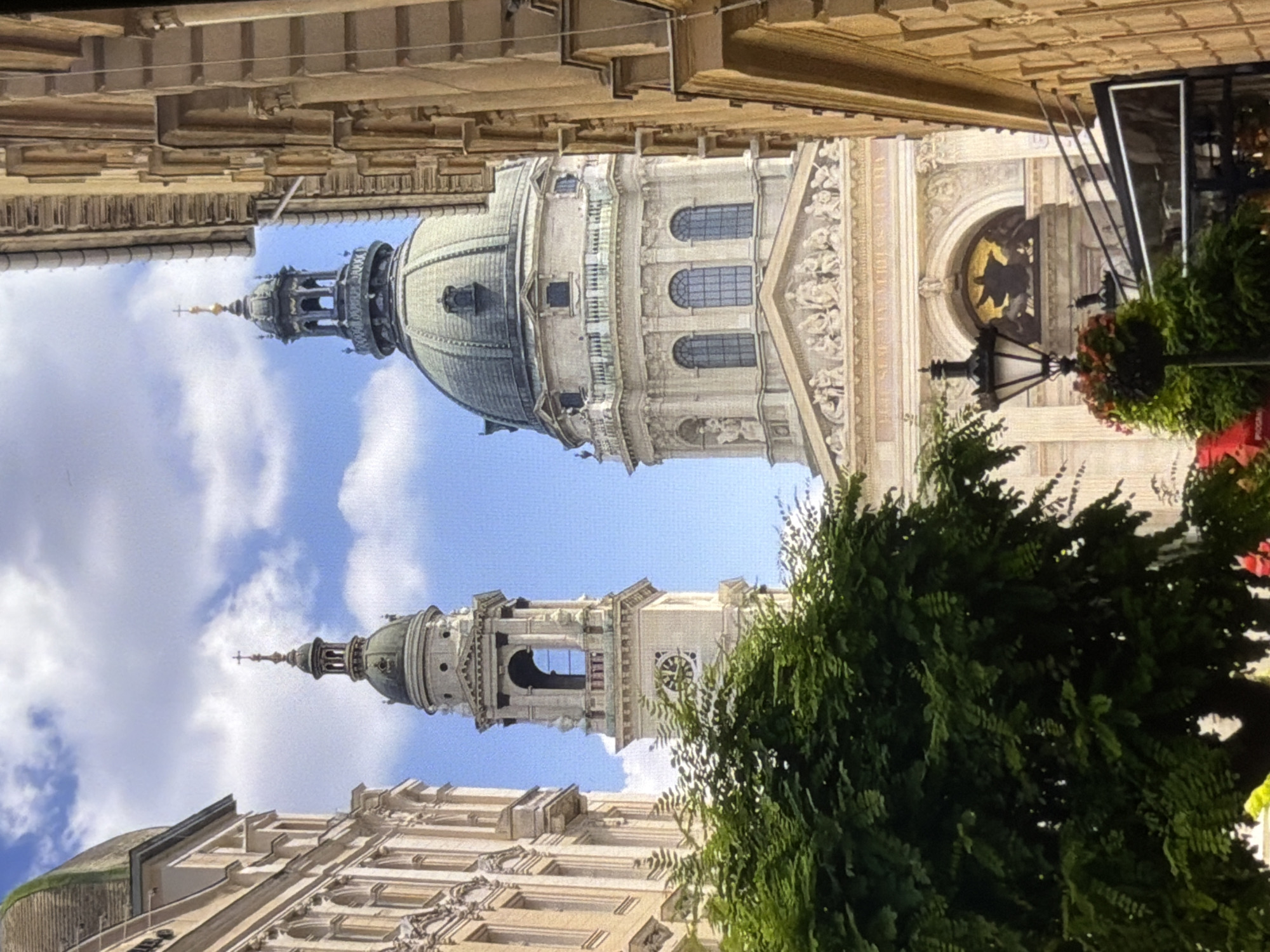 The image shows a low-angle, vertical view of an ornate architectural structure, likely a cathedral or church, with a prominent dome and a tall, slender tower. The building is made of light-colored stone and features intricate carvings and arched windows. The dome is topped with a cross, and the tower has a clock face visible. Lush green foliage is visible in the foreground, partially obscuring the lower parts of the building and adding a natural element to the scene. The sky above is a clear blue with scattered white clouds, suggesting it is daytime and the weather is pleasant. The architecture suggests a European location, possibly a historical city. There is no discernible activity or people in the image. The overall impression is one of grandeur and historical significance.