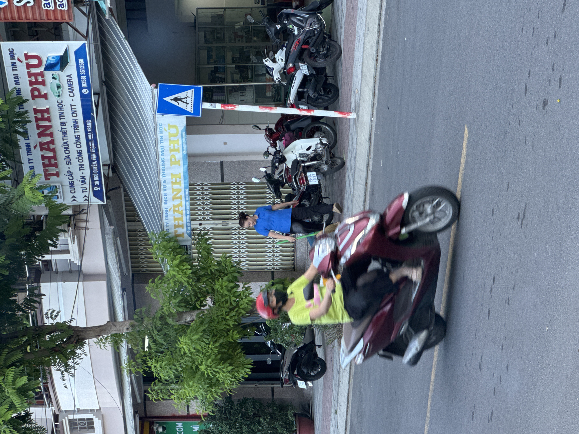 The image depicts a street scene in Nha Trang, Vietnam. In the foreground, a person wearing a bright green t-shirt, black pants, and a red helmet is riding a dark red scooter. They are wearing a black mask that covers their nose and mouth. The scooter is moving, as indicated by motion blur on the wheels and the vehicle itself. To the right of the scooter rider, a woman in a blue shirt and black pants is walking and appears to be holding a broom or similar cleaning implement. Behind the woman, several motorcycles are parked on the sidewalk.
The background features a building with awnings and signs. One prominent sign, in Vietnamese, displays "THÀNH PHÚ" in large red lettering and includes an image of a laptop and a camera. Another smaller blue sign indicates a pedestrian crossing. Trees with lush green foliage are visible on the left side of the image, obscuring parts of the building. The street surface is asphalt with a yellow line painted on it. The overall lighting suggests it is daytime.