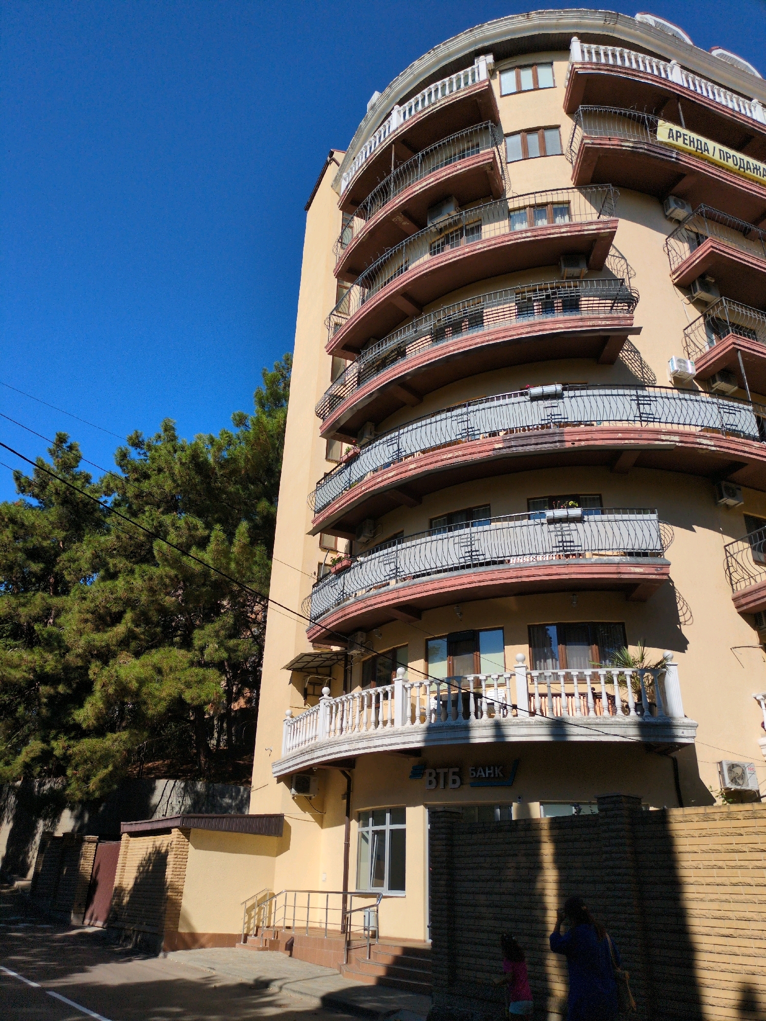 The image shows a light yellow, curved building with multiple balconies. The building has a light brown trim around the balconies and white railings. There is a banner with Russian text that appears to say "АРЕНДА / ПРОДАЖА", which translates to "RENT / SALE". Below the balconies, there is a sign that says "ВТБ БАНК". The building is surrounded by trees on the left and a brick wall on the right. The sky is a clear blue. There are two people visible in the lower right corner of the image: one adult wearing a blue jacket and carrying a bag, and a child wearing a pink shirt. They are standing near the brick wall. The scene appears to be a sunny day in an urban environment.