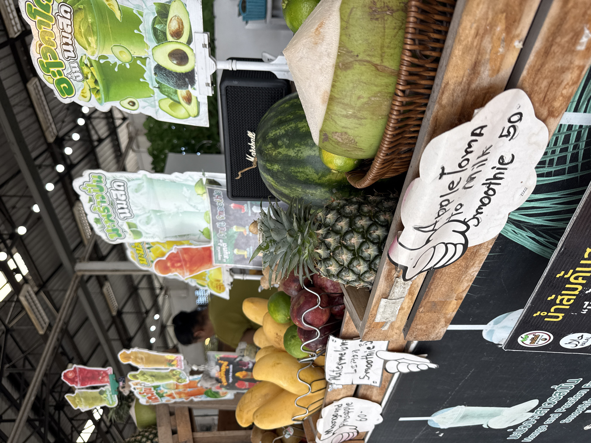 This image captures a vibrant fruit and smoothie stand at the Fisherman's Village Walking Street in Baan Bo Phut, Koh Samui, Thailand. In the foreground, a variety of fresh fruits are displayed, including a pineapple, mangoes, limes, and a whole coconut. Behind the fruits, a large watermelon is visible, along with a Marshall speaker. Several signs advertising different smoothie flavors, such as "Apple Toma to milk smoothie," "Watermelon Lemon Smoothie," and "Mango and Pineapple," are placed around the stall. In the background, more signs featuring colorful fruit illustrations are displayed under a covered market roof. A person wearing a green shirt is partially visible in the background, attending to customers or preparing drinks. The overall scene depicts a lively outdoor market atmosphere with a focus on fresh produce and beverages.