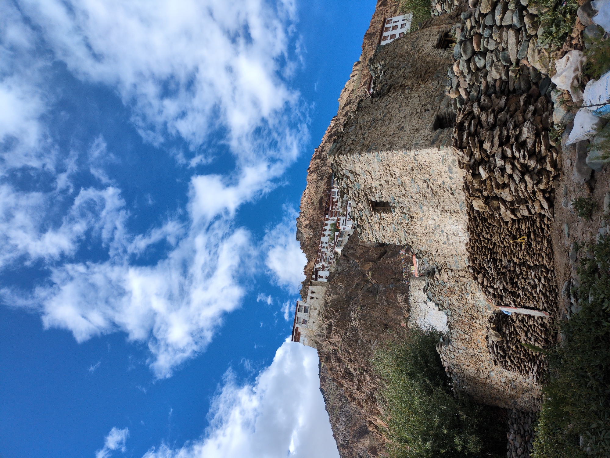 This image captures a dramatic scene of a monastery built precariously into a rocky cliff face. The monastery's white buildings with red trim stand out against the rough, brown stone of the mountain. The structures appear to be multi-storied and cling to the sheer rock, suggesting a remarkable feat of engineering and a deep connection to the natural environment.
Below the main monastery, a section of the cliff is reinforced with a wall constructed of numerous stones, creating a textured and robust barrier. Some of the stones are quite large, while others are smaller and more rounded, indicating a careful selection and placement. Green foliage, including trees and bushes, grows around the base of the cliff and along the edges of the constructed areas, adding a touch of life to the arid landscape.
The sky above is a vibrant blue, filled with fluffy white clouds that are dramatically illuminated by sunlight, suggesting a bright and clear day. The angle of the light indicates it could be late morning or early afternoon. The overall impression is one of resilience, devotion, and a harmonious coexistence between human habitation and the rugged terrain.
The location is identified as Yulang, India, which is known for its mountainous landscape and spiritual sites. There is no visible text within the image.