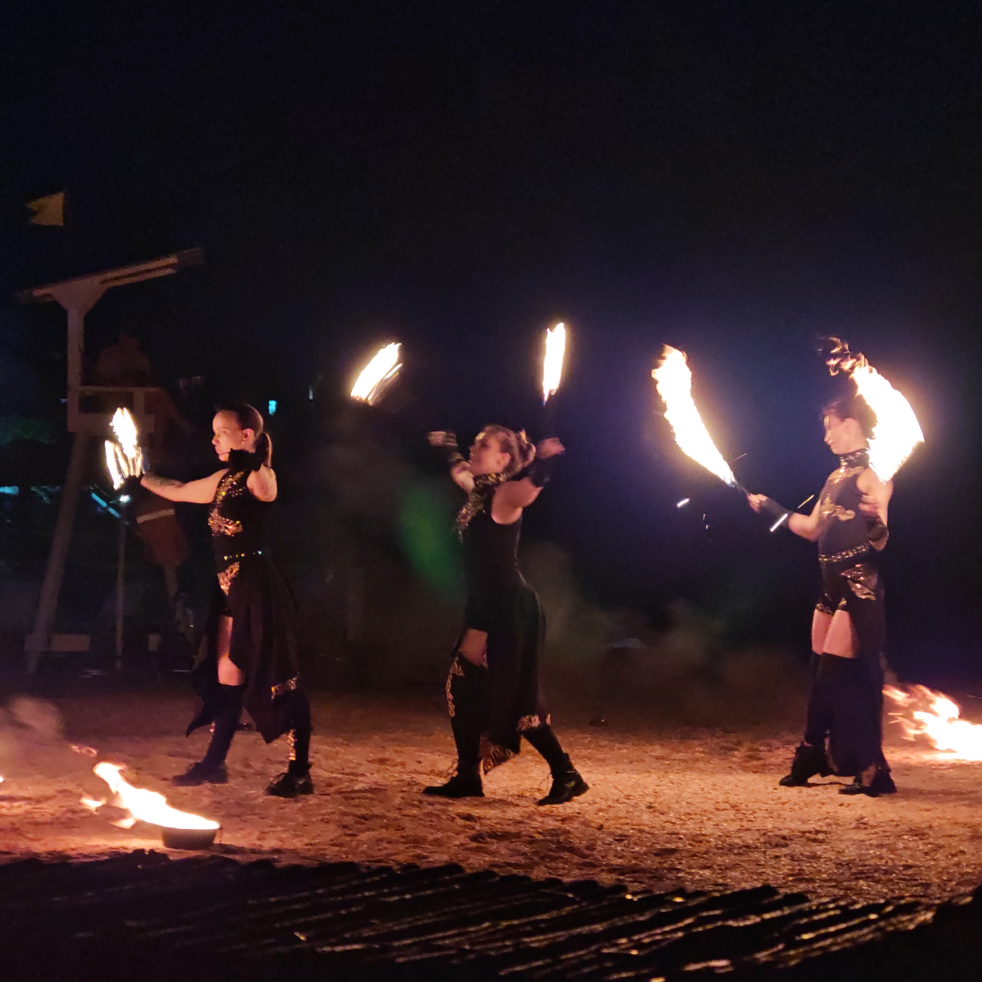 Three women are performing fire dancing in a dark, outdoor setting, likely a public event or festival. The ground appears to be a sandy or dirt path, with some dry brush or straw visible in the foreground. The women are dressed in dark, ornate costumes with some metallic embellishments. They are all holding and manipulating fire, either with torches or by spinning fire props. The flames are bright and cast a warm glow on their faces and costumes, as well as the surrounding area. In the background, there is a wooden structure, possibly a stage or scaffolding, with a yellow flag attached to it. The overall atmosphere is dramatic and energetic, characteristic of a fire performance.