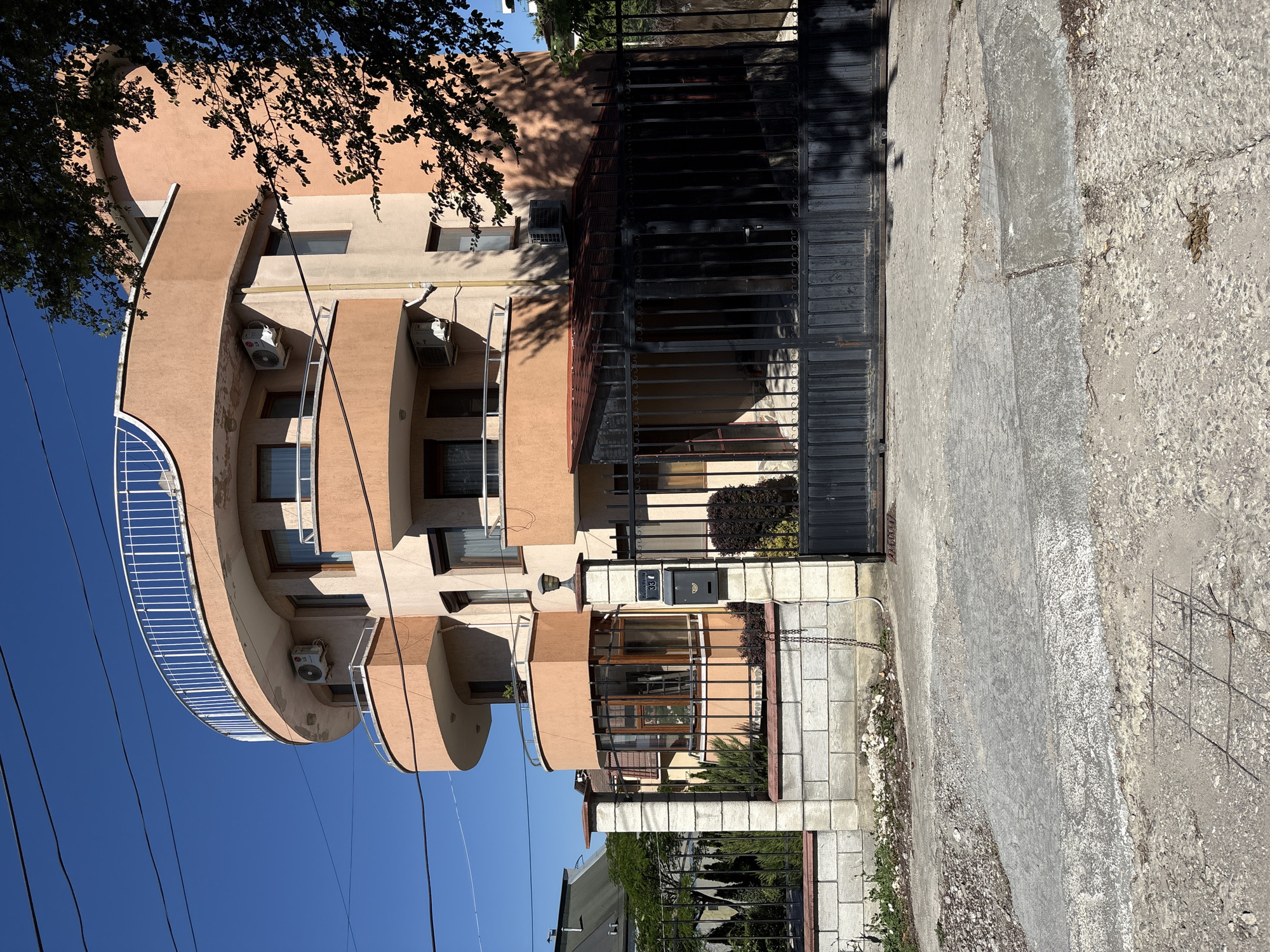 A three-story residential building with a curved, light-orange facade and multiple balconies is shown from the street in Varna, Bulgaria. The building has a clear blue sky behind it and some tree branches obscure the upper right portion of the facade. A black metal gate and fence, constructed with stone pillars, encloses the property. The gate is closed, and a mailbox is affixed to one of the gateposts. The street in front of the property is paved with cracked concrete and gravel.