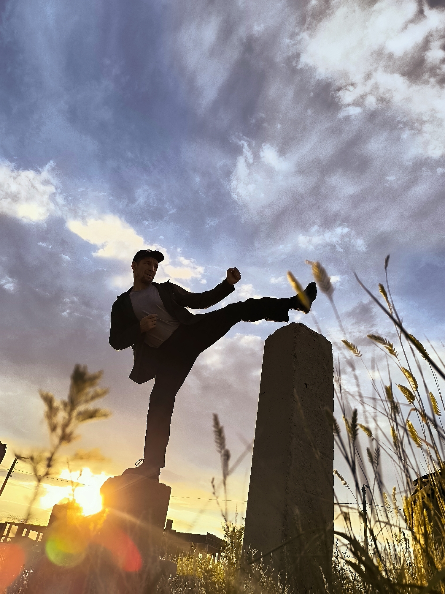 A man is silhouetted against a dramatic sky, performing a martial arts kick. He stands on a concrete pillar, with his right leg extended forward in a dynamic pose. He wears dark pants, a grey t-shirt, a dark jacket, and a baseball cap. His left leg is bent, providing balance. His right fist is clenched, ready for action.
The scene is set at sunset, with the low sun casting a warm, golden glow and creating lens flare effects. The sky is filled with a mix of dark, ominous clouds and brighter, lighter patches, suggesting dynamic weather. Tall, dry grasses are in the foreground and to the right, adding a natural, slightly wild element to the urban setting. In the background, hints of buildings and utility poles can be seen.
The man appears focused and powerful, mid-action in his martial arts move. The overall mood is one of energy and determination, set against the backdrop of a striking natural phenomenon. There is no visible text in the image. The location context provided places this scene in the Qarağandı city administration in Kazakhstan.