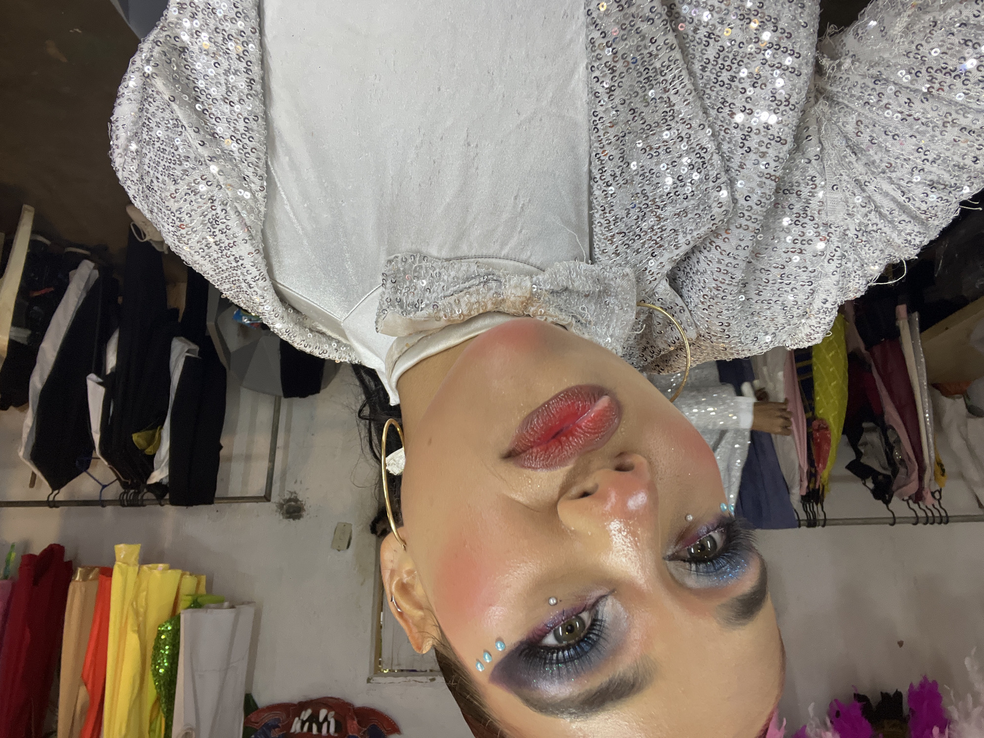 A close-up, medium-shot selfie of a woman with dramatic makeup is taken from a low angle in what appears to be a backstage area. The woman is looking directly at the camera. She has bright blue eyeshadow with glitter, false eyelashes, and sparkly embellishments on her cheek. Her lips are painted a glossy red, and she is wearing large hoop earrings and a sparkly silver jacket over a white garment. Behind her, racks of colorful costumes and clothing are visible, along with a white wall. The lighting is somewhat harsh.