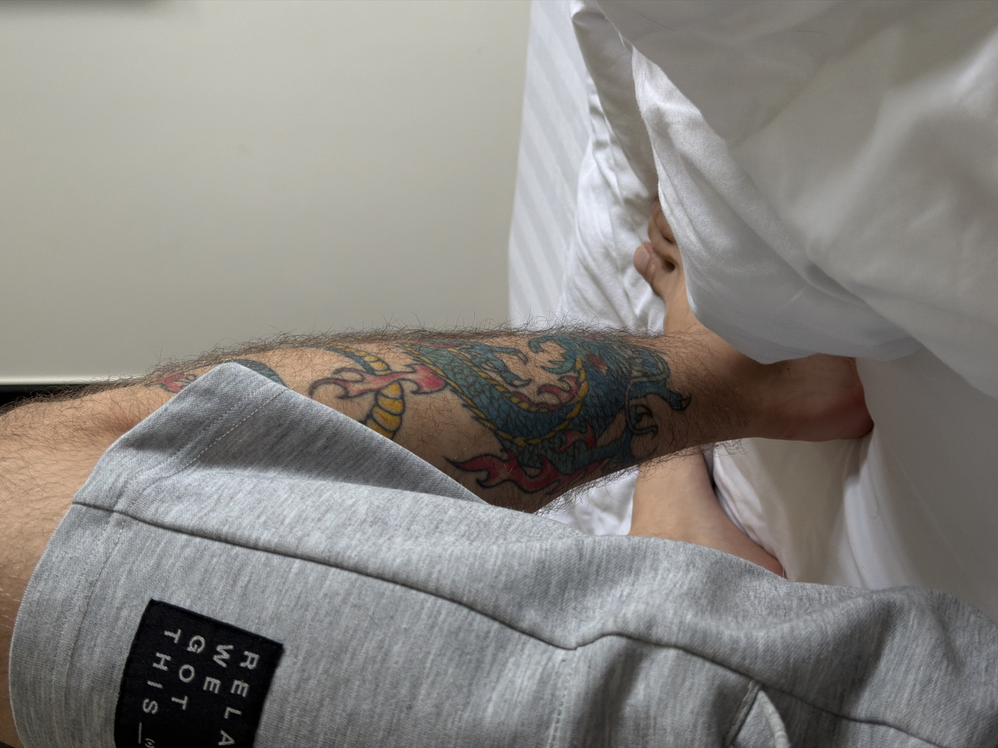 A first-person perspective image shows a man lying in bed with a visible tattoo of a dragon on his right calf. He is wearing gray sweatpants. The bed has white sheets and a comforter. The background includes a light gray wall. There is no information to place the scene in Đà Nẵng, Vietnam.