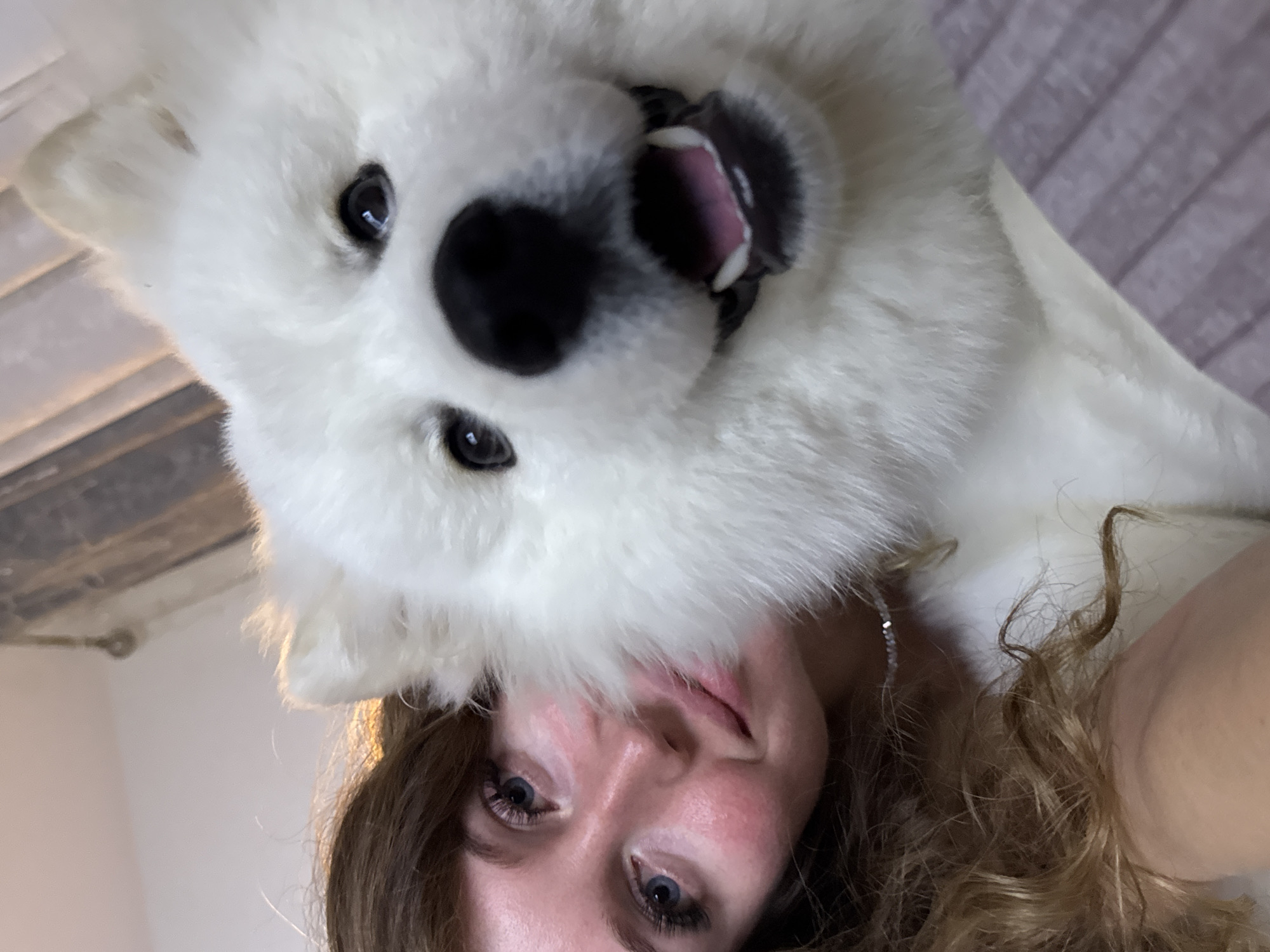 A close-up selfie shows a young woman with long, wavy brown hair and blue eyes. She is wearing makeup, including eyeliner and mascara, and has blush on her cheeks. Her left shoulder and upper chest are visible, and she is wearing a delicate necklace. Partially obscuring the woman is a large, fluffy white dog, likely a Samoyed, with its head tilted towards the camera. The dog has dark eyes and an open mouth, appearing to smile. The background is blurred but suggests an indoor setting with warm lighting and a hint of furniture. The scene is likely a private residence. There is no identifiable information indicating the specific location in Chaikovsky, Russia.