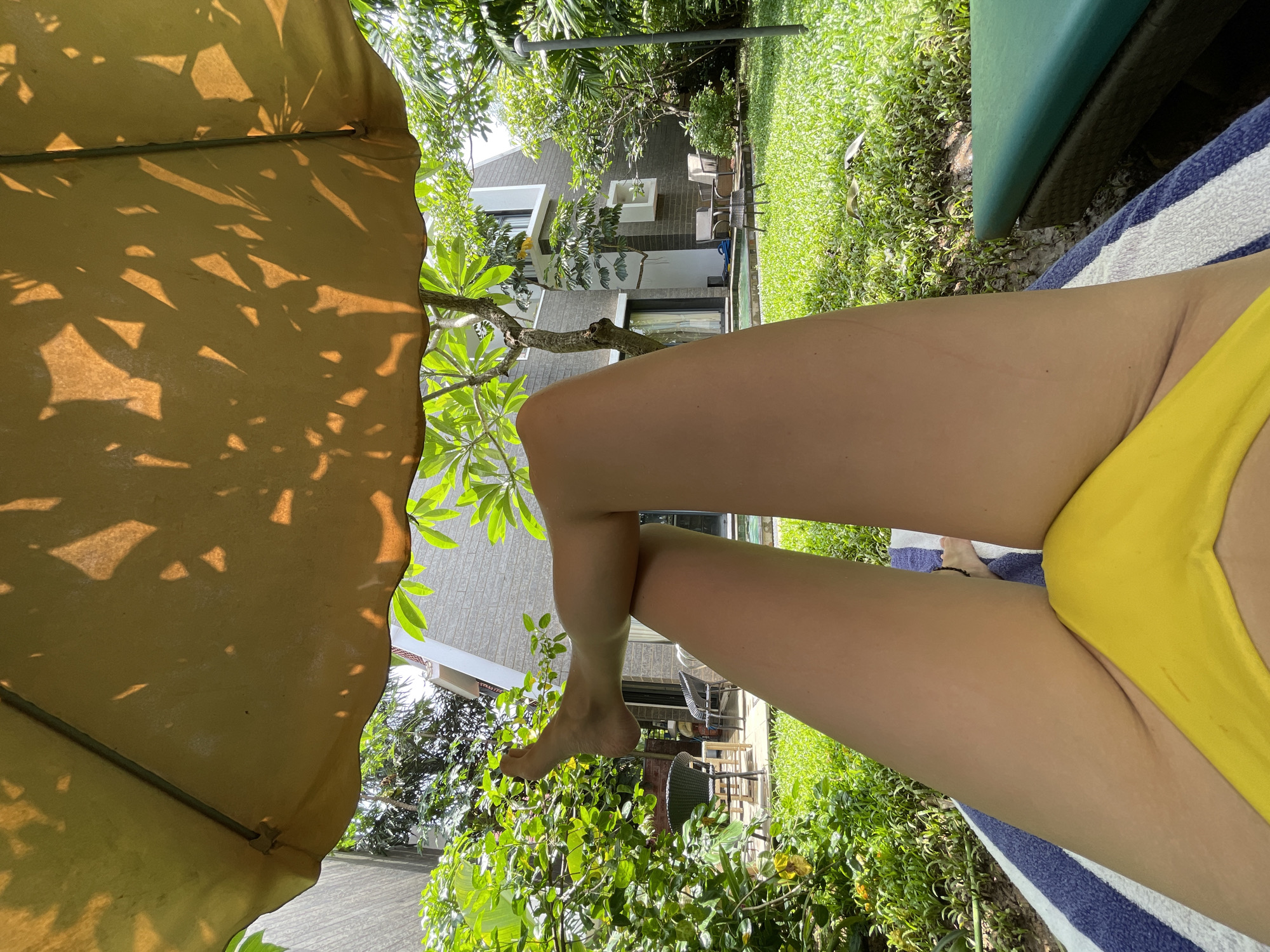 A person is lying on a blue and white striped towel on a lounge chair in what appears to be a backyard or resort setting. The person is wearing a bright yellow bikini bottom. Their legs are extended upwards, partially obscuring the view of the surroundings. In the background, there is a well-maintained lawn, lush green trees and plants, and a modern building with grey brick and white accents. There are also some outdoor chairs and a small table visible. A large, yellow umbrella with leaf shadows cast on it provides shade. The scene suggests a relaxing day outdoors, likely in a warm climate. The location is specified as Badem, India.