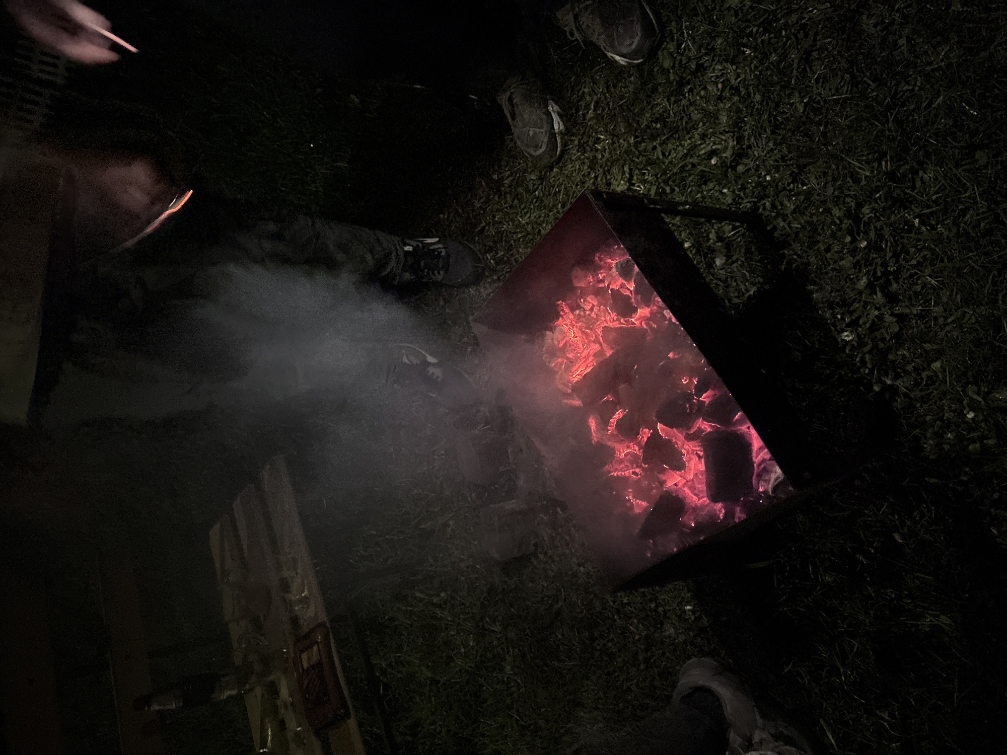 The image shows a dark outdoor scene, likely taken at night in Пярэжырскі сельскі Савет, Belarus. A metal fire pit containing hot, glowing embers is prominently featured. Smoke is visible rising from the fire pit. At least three people are present, indicated by their feet and legs, but their faces are not visible. The ground appears to be covered in grass. One hand appears to be holding an object, possibly a stick or sparkler, near the smoke. A wooden structure or object is also visible to the left of the fire pit.