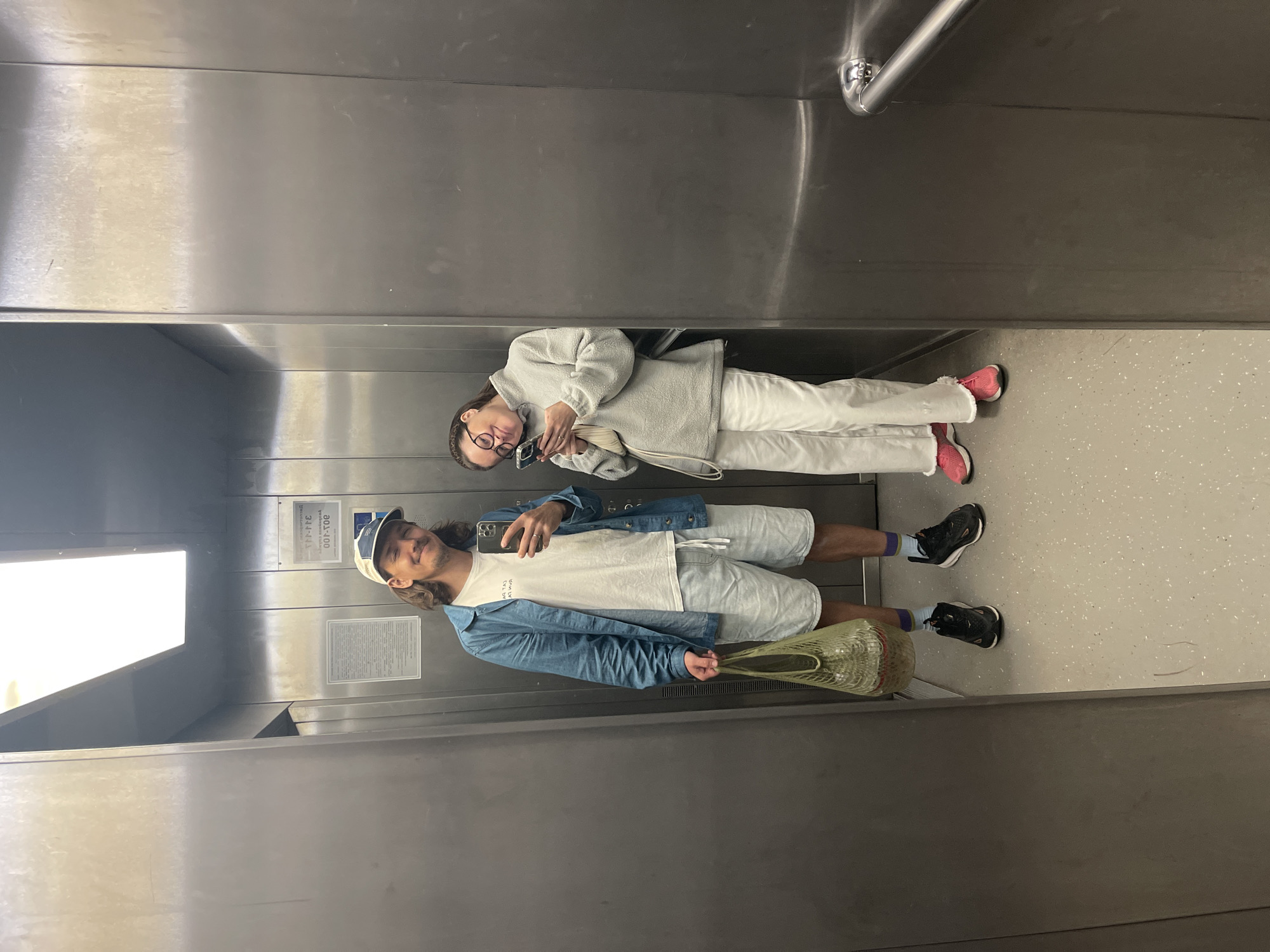 In an elevator with brushed metal walls, a man and a woman pose for a selfie. The man, on the left, wears a blue denim jacket over a white t-shirt, light wash denim shorts, and black sneakers with blue and purple striped socks. He holds a green mesh bag in his left hand and his phone in his right. He has long brown hair and a beard, and wears a beige baseball cap. The woman, on the right, is dressed in a light grey fleece sweatshirt, white pants, and bright pink sneakers. She holds her phone and smiles. The elevator floor is light grey with speckled dark grey flecks. A white sign with Cyrillic text is affixed to the wall behind the man, and a silver handrail is visible on the right wall.