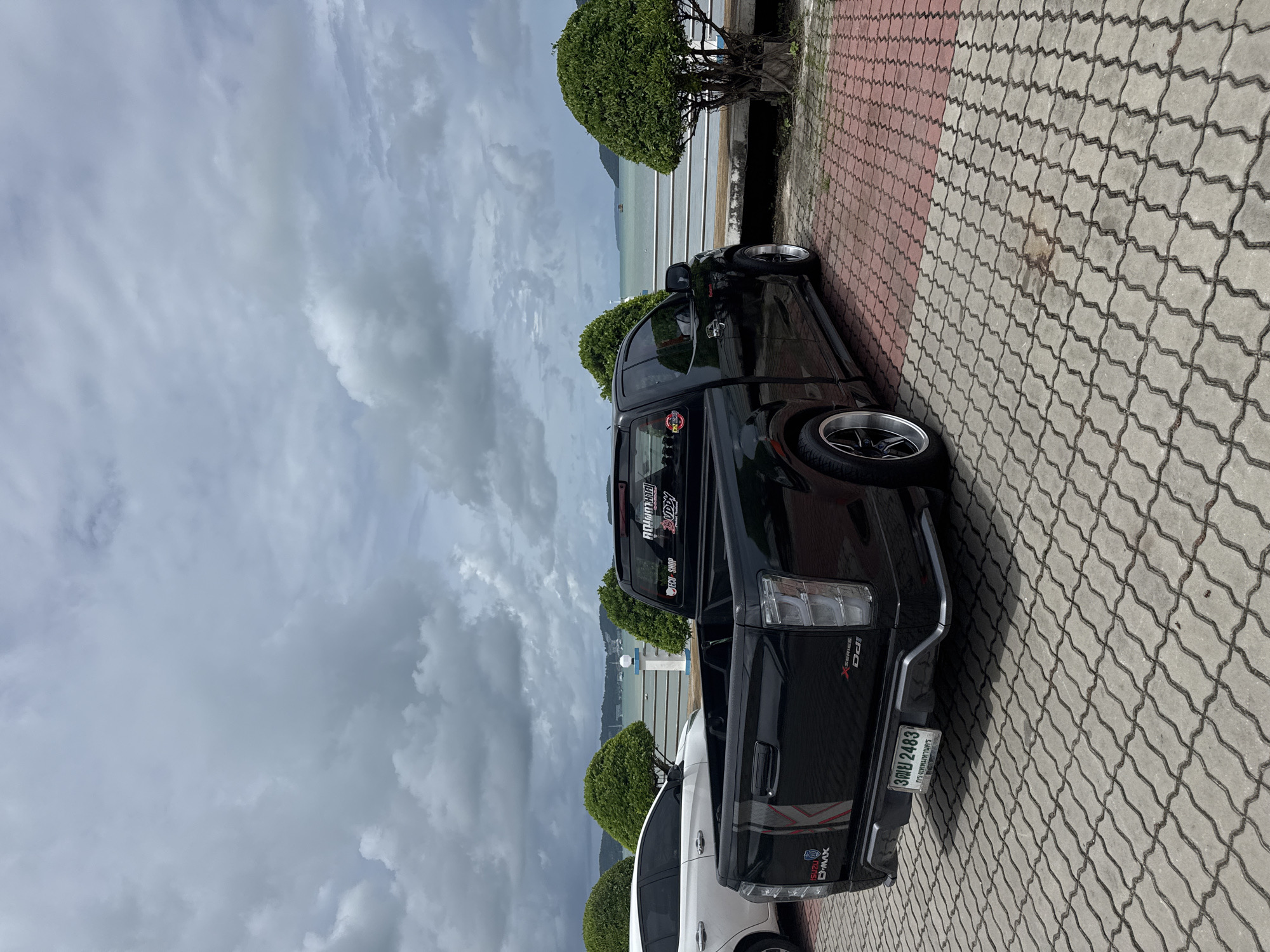 A black Isuzu D-Max pickup truck is parked on a paved area near the coast in Rawai, Thailand. The truck is facing away from the camera and is positioned on the right side of the image. It has tinted windows and custom rims. To the left of the truck, a white car is partially visible.
The environment includes a body of water, likely the Andaman Sea, with a cloudy sky overhead. A railing separates the paved area from the water. Two well-maintained, rounded bushes are placed symmetrically on either side of the truck, framing the scene. The paving stones are a mixture of red and gray interlocking bricks. In the distance, islands or the mainland coastline can be seen. There are no people visible in the image. The overall atmosphere is calm and overcast.