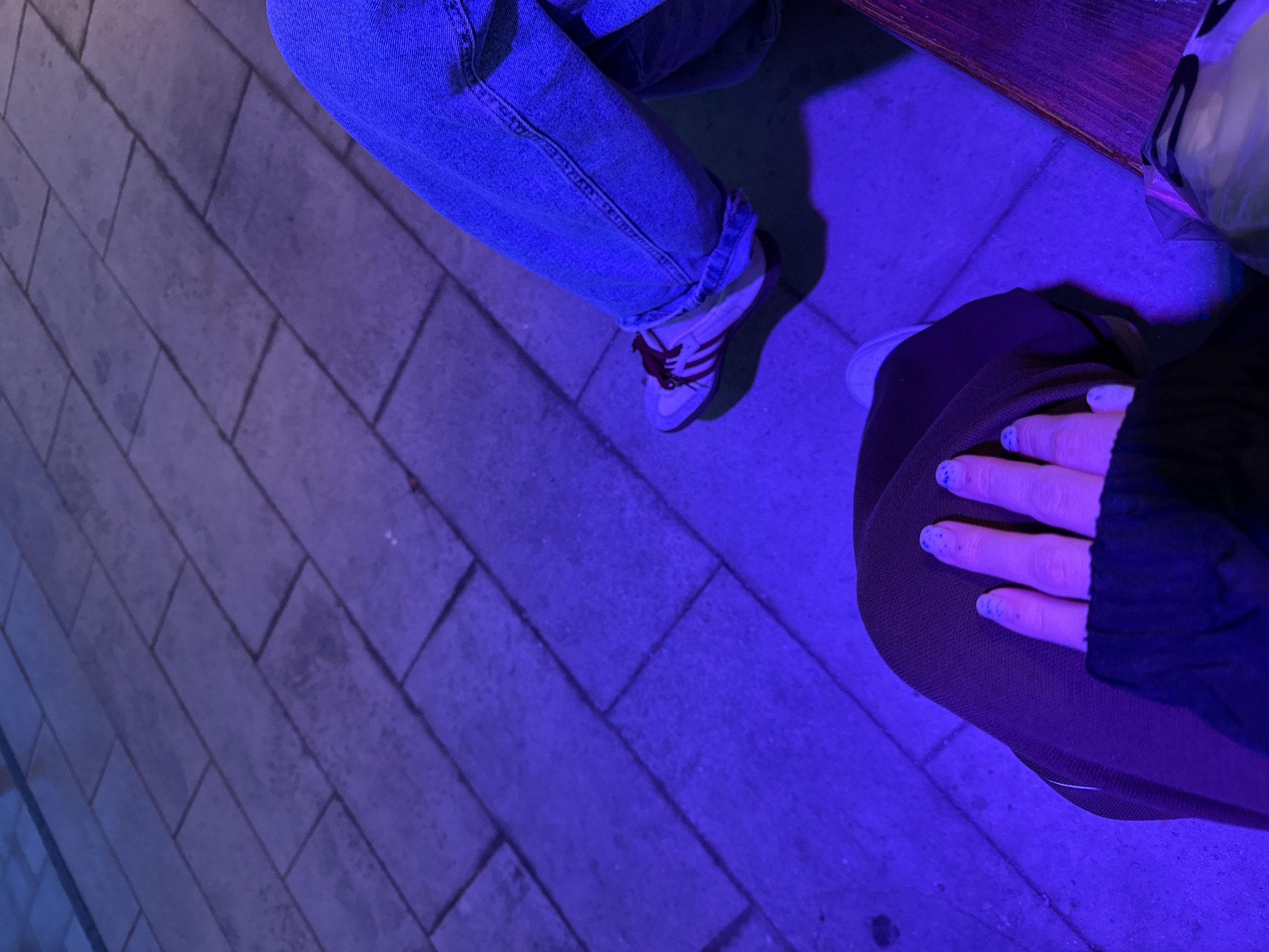 The image shows two people sitting outdoors at night in Rostov-on-Don, Russia. The ground is paved with rectangular stones. One person is wearing blue jeans and white sneakers with red stripes. The second person is wearing brown pants, a black jacket, and has light skin with blue nail polish on their fingernails. Their hand rests on their thigh. The scene is illuminated by a strong blue light. Part of a wooden table and a white bag are visible in the top right of the frame.