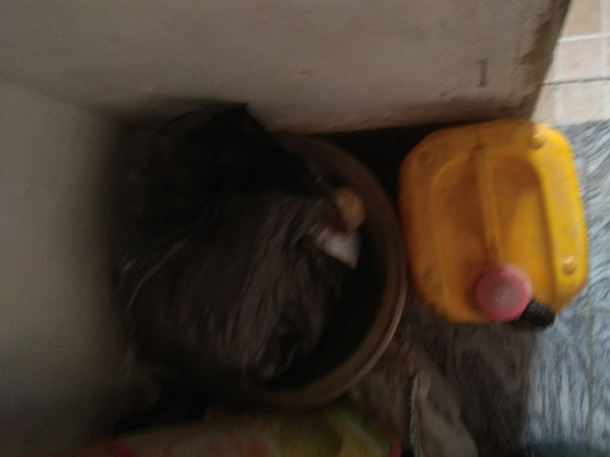 The image shows a top-down view of a corner in what appears to be an indoor setting, possibly a storage area or a less-trafficked part of a building. The scene contains several objects. To the left, there is a large, dark-colored bag, possibly a trash bag, resting inside a shallow, round container. The bag is full and appears to be made of a dark plastic material. To the right of the bag is a bright yellow plastic jerrycan. It has a handle on top and a pink cap or stopper on its side. The jerrycan is positioned on a grey, textured surface, which might be a mat or a type of flooring. Behind the objects, a beige-colored wall or surface forms the background, meeting a lighter-colored surface at an angle, suggesting a corner of a room. The lighting is dim, casting some shadows and making some details blurry. The overall impression is one of everyday storage or a utility area, with no specific activity or event clearly depicted. The location context is Jalingo, Nigeria, but there are no explicit visual cues within the image to confirm this. No text is visible in the image.