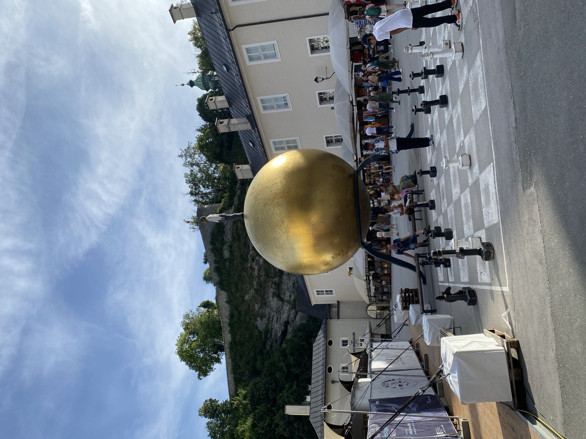 The image depicts the "Sphaera" sculpture by Stephan Balkenhol, a large golden sphere topped with a figure of a man, located in the Residenzplatz square in Salzburg, Austria. In front of the sculpture, a giant chessboard is set up with large black and white chess pieces. Several people are visible in the square, some observing the sculpture, others walking around or playing chess. The background shows traditional Salzburg architecture, including buildings with light-colored facades and a green-domed church tower, with a hillside covered in greenery and a wall in the upper part of the frame. The sky is blue with some wispy clouds. The scene appears to be set during daytime, likely a warm season due to the attire of the people.