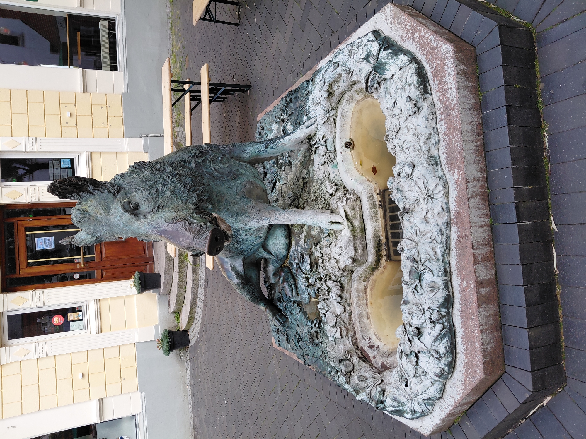 The image shows a bronze sculpture of a wild boar in the city of Holstebro, Denmark. The sculpture is part of a fountain with two basins filled with water. The boar is depicted in a reclining pose, with its head raised and mouth slightly open. The sculpture is surrounded by a decorative base with floral motifs. The fountain is located in a paved outdoor area, possibly a public square or street, with a building in the background that appears to be a shop or restaurant with a wooden door and windows. There are also some wooden picnic tables visible in the background. The paving is made of dark interlocking bricks. The overall impression is of a charming and artistic public installation.
