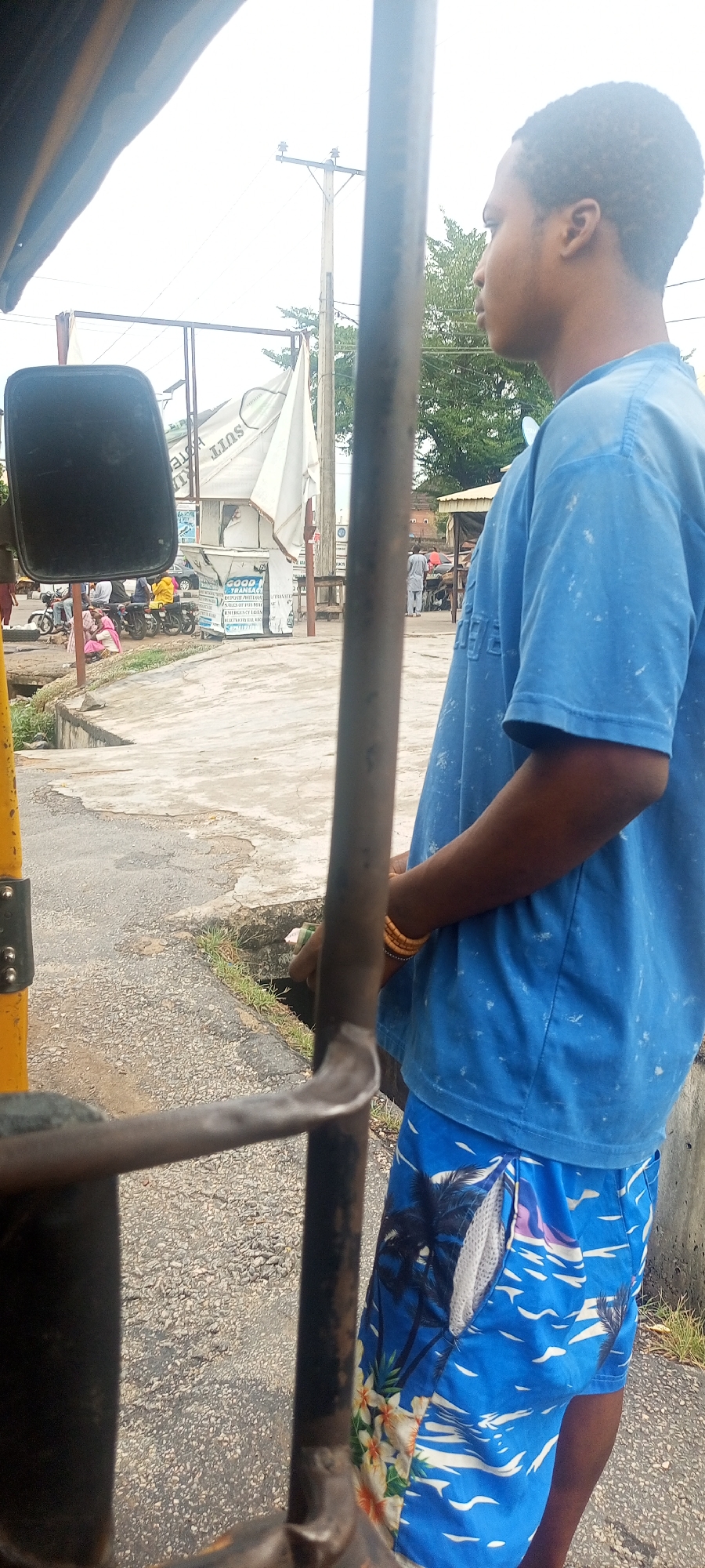 An image shows a person in Adankolo, Nigeria, seen from within a vehicle, possibly a "Keke Napep" or auto-rickshaw. The person is wearing a light blue T-shirt and patterned blue shorts with palm trees and flowers. The vehicle's frame and a side mirror are visible in the foreground. In the background, there is a street scene with buildings, signs, and other people. There appears to be a makeshift market area or small shops in the background.