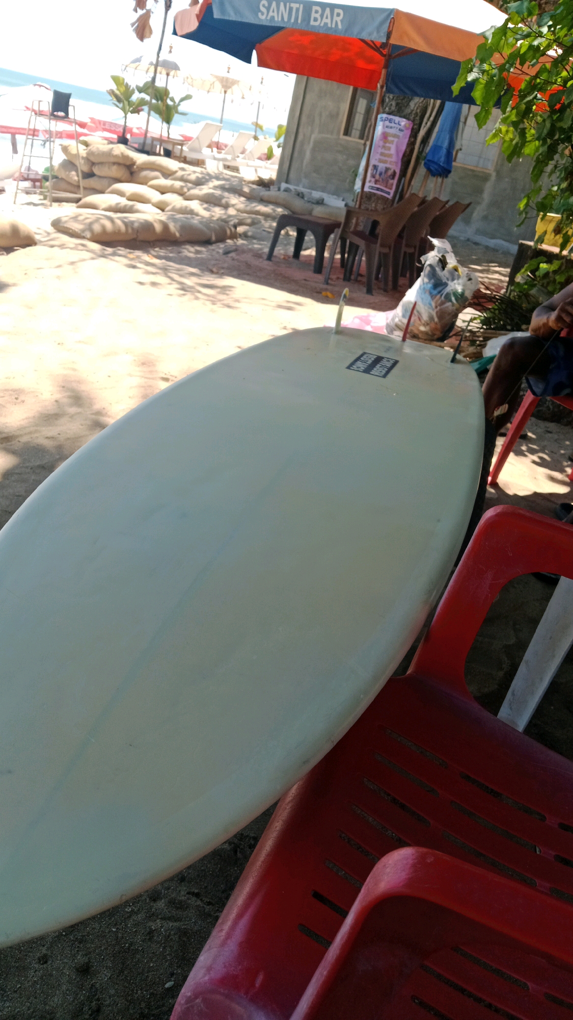 An outdoor, low-angle shot shows a light green surfboard resting on a sandy surface next to two red chairs. The surfboard is the primary subject, occupying the majority of the foreground. Behind the surfboard and chairs, a beach scene unfolds, featuring sandbags, lounge chairs, and umbrellas. A building with a sign that reads "SANTI BAR" is visible in the middle ground, suggesting the location is a beachside establishment in Kuta, Indonesia. The atmosphere appears casual and relaxed, with the bright sunlight casting shadows and highlighting the textures of the sand and the surfboard.