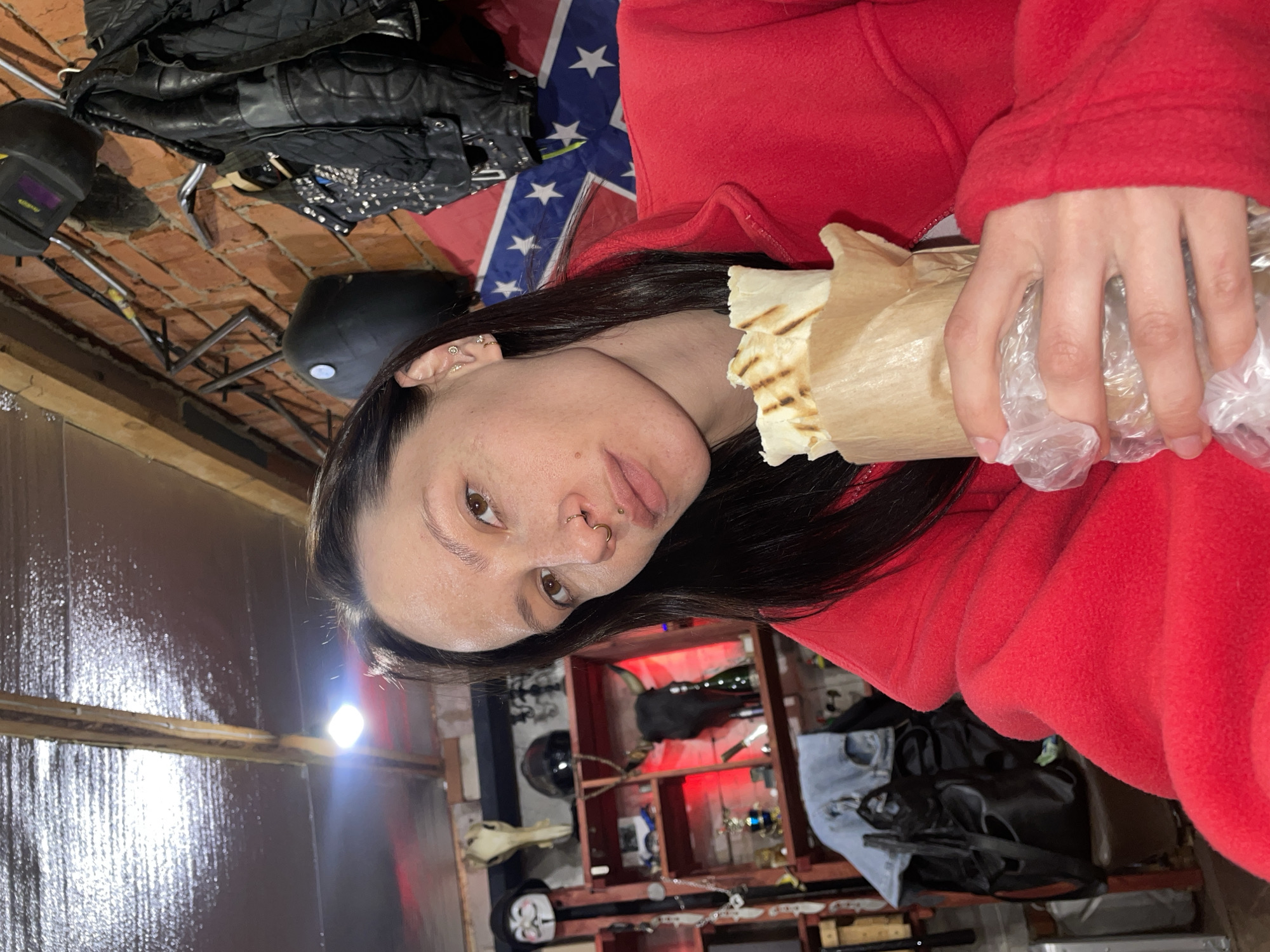 A woman with dark hair, wearing a red fleece jacket, holds a wrapped food item (likely a kebab) in her hands. She has facial piercings. Behind her, a Confederate flag is visible, along with a black leather jacket. In the background are various objects on shelves and a brick wall. The location is unclear. There is no information to suggest this location is in Oktyabr'skii, Russia.