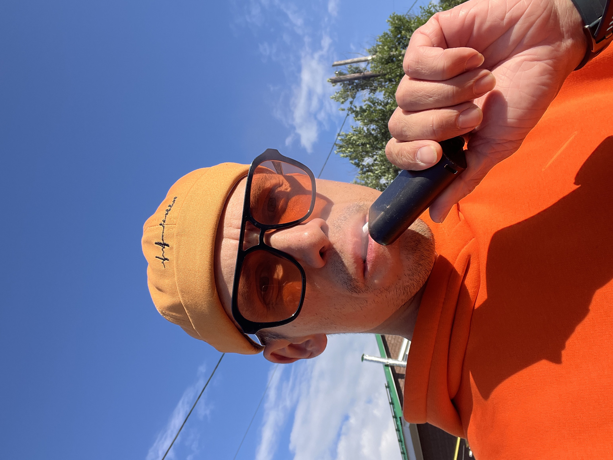 A man, seen from a low angle, is partially captured against a bright blue sky with scattered white clouds. He wears an orange beanie with some black embroidery on the left side and orange-tinted sunglasses with a black frame. His face is angled upwards, revealing stubble, and he has a black object, possibly a vape or electronic device, positioned at his mouth. A man's hand, wearing a dark wristband or watch, holds this object. He is dressed in an orange hoodie or sweatshirt. In the background, green trees and utility poles with wires are visible. The lighting suggests it is daytime, under sunny weather conditions. The overall setting is outdoors, likely in an urban or suburban area, specifically in Chaikovsky, Russia. No specific emotions are discernible, and the embroidered text on the beanie is not clearly legible.