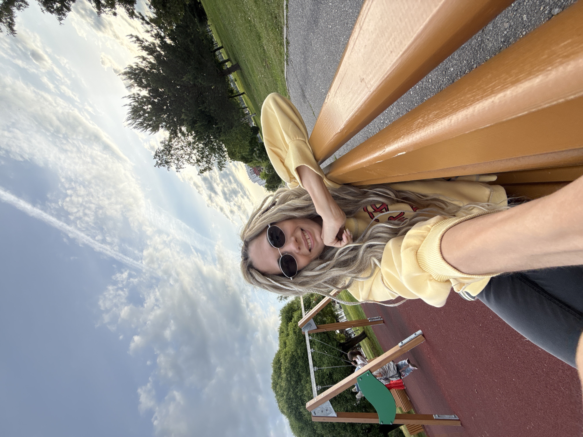 The image, oriented diagonally, features a young woman taking a selfie in what appears to be a park or recreational area in Заволжье, Russia. She is seen from a low angle, resting on a brown slatted bench, smiling at the camera. She wears round dark sunglasses and has long, light-colored hair styled in dreadlocks or braids. Her attire includes a yellow long-sleeved top, possibly a hoodie, which has an indistinct red and white graphic on the front, and dark pants.
The background reveals a green landscape with various trees, a grassy expanse, and a paved path. Further in the background, a children's playground is visible, featuring a swing set structure and a reddish ground surface. The sky is partly cloudy with some clear blue patches and faint contrails, suggesting a bright late afternoon or early evening setting. The woman's smile conveys a happy and relaxed mood. No other legible text is visible in the scene.