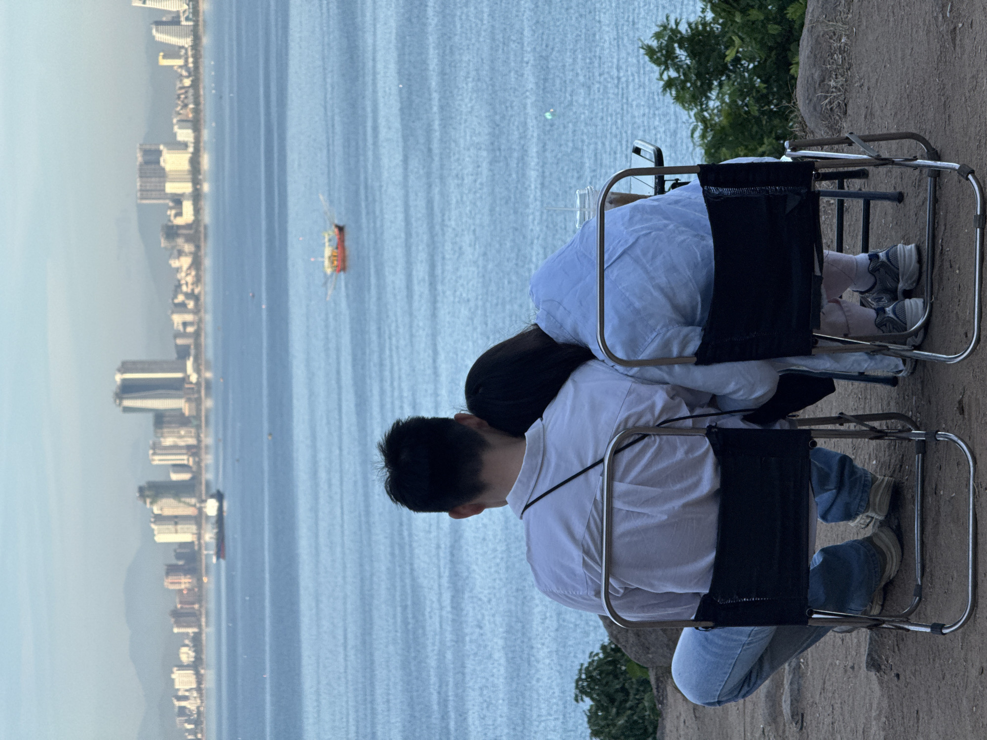 The image shows a couple sitting on chairs overlooking the city of Da Nang, Vietnam. The city skyline, with its numerous buildings and a distant mountain range, is visible across the ocean. The water is calm with a small red boat in the distance, and a larger ship closer to the city. The couple, seen from behind, are embracing, with one person resting their head on the other's shoulder. They are seated on foldable chairs on a rocky, elevated ground with some vegetation. The lighting suggests it is either early morning or late afternoon.