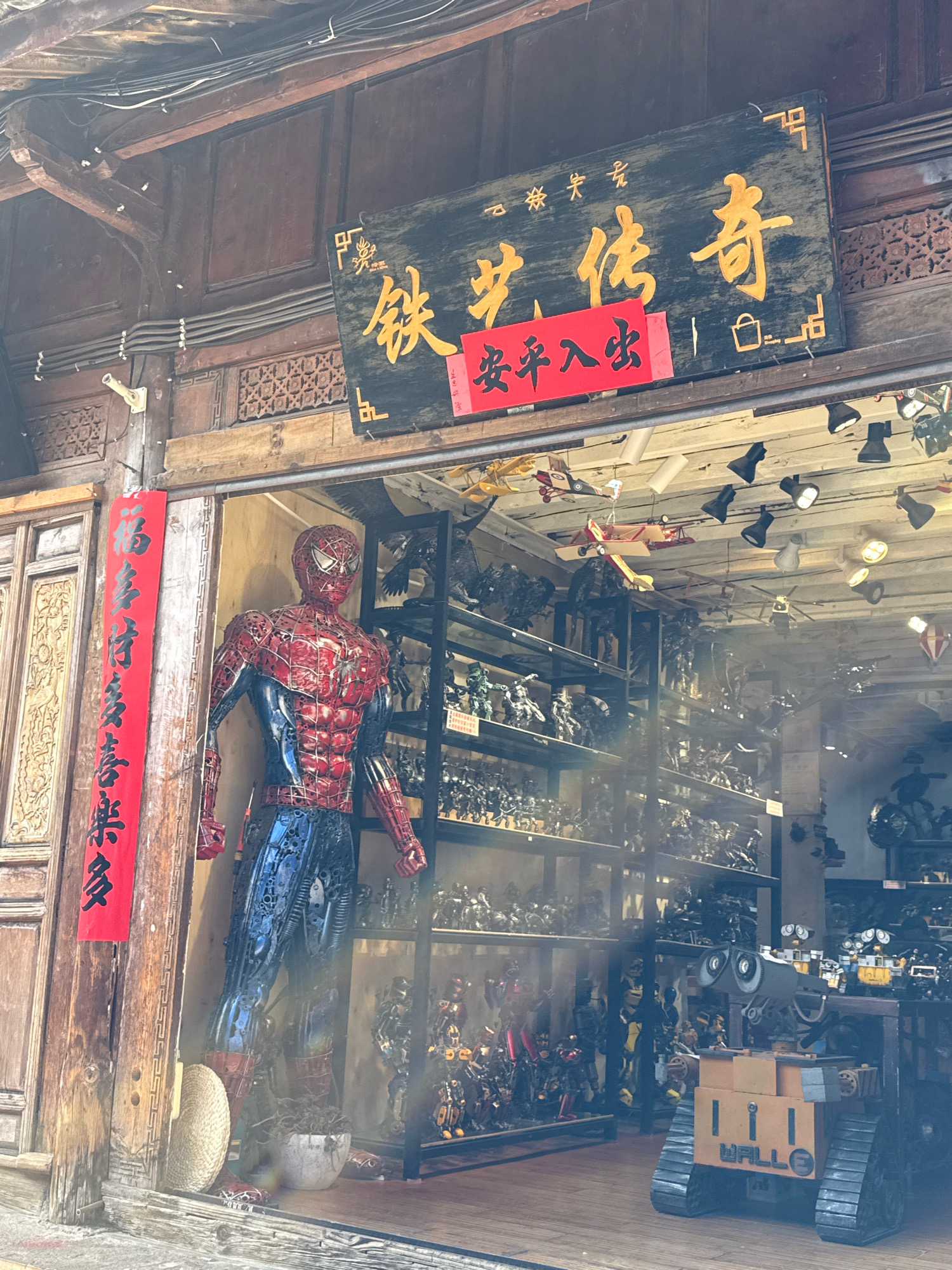This is a photograph of the exterior of a shop in Gucheng District, China. The shop's facade is made of traditional wooden architecture. A large, metallic sculpture of Spiderman stands on the left side of the entrance. To the right of Spiderman, a shelf unit displays numerous small action figures and models of robots and vehicles. In the foreground, on the right, a recognizable WALL-E robot model is visible. Above the entrance, a large wooden sign with golden Chinese characters is displayed, and below it, a red banner with black characters is hung. The interior of the shop appears to be filled with more shelves of merchandise, including model airplanes suspended from the ceiling. The lighting suggests it is daytime.