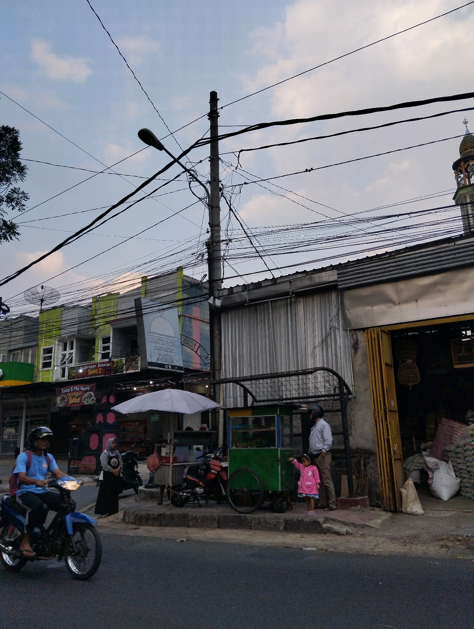 The image depicts a street scene in Salatiga, Indonesia, with a focus on commercial activity and daily life. In the foreground, a young person rides a blue motorcycle eastward on a paved road. The rider is wearing a blue t-shirt, dark pants, and a black helmet. Behind the rider, on the sidewalk, a woman in a black headscarf and dark clothing stands near a parked motorcycle. Further down the sidewalk, a child in a pink jacket points towards a green food cart, beside which stands a person in a light-colored shirt and brown pants, possibly a vendor. The food cart is positioned next to a stall with various goods, including large bags of what appear to be rice or grain.
The background features a row of buildings with a mix of architectural styles. One building, painted in shades of green and white, has signage indicating it houses a mosque and "Bakso & Mie Ayam". Another building has a large, faded mural of a mosque. Overhead, a dense network of electrical wires crisscrosses the sky, supported by poles. A mosque minaret with a golden dome is visible in the far right background. The sky is cloudy, suggesting either early morning or late afternoon light. The overall impression is of a bustling urban environment with a strong presence of small businesses and residential areas.