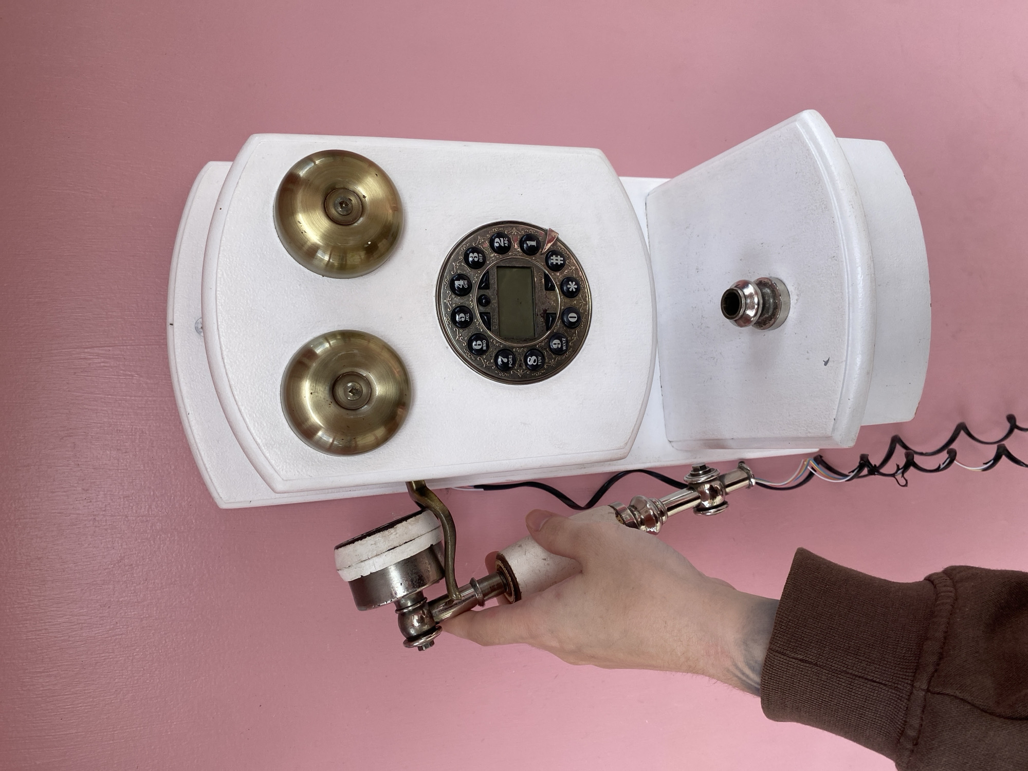 A person's hand is holding an antique white rotary phone against a bright pink wall. The phone is mounted on the wall and has two brass circular pieces that resemble eyes, a circular dial with numbers and symbols, and a handset. The handset is currently being held by the person's hand. Below the dial is a shelf with a small metal piece attached to it. The image is a close-up shot, focusing on the phone and the hand interacting with it. No specific location in Izhevsk, Russia can be determined from this image.