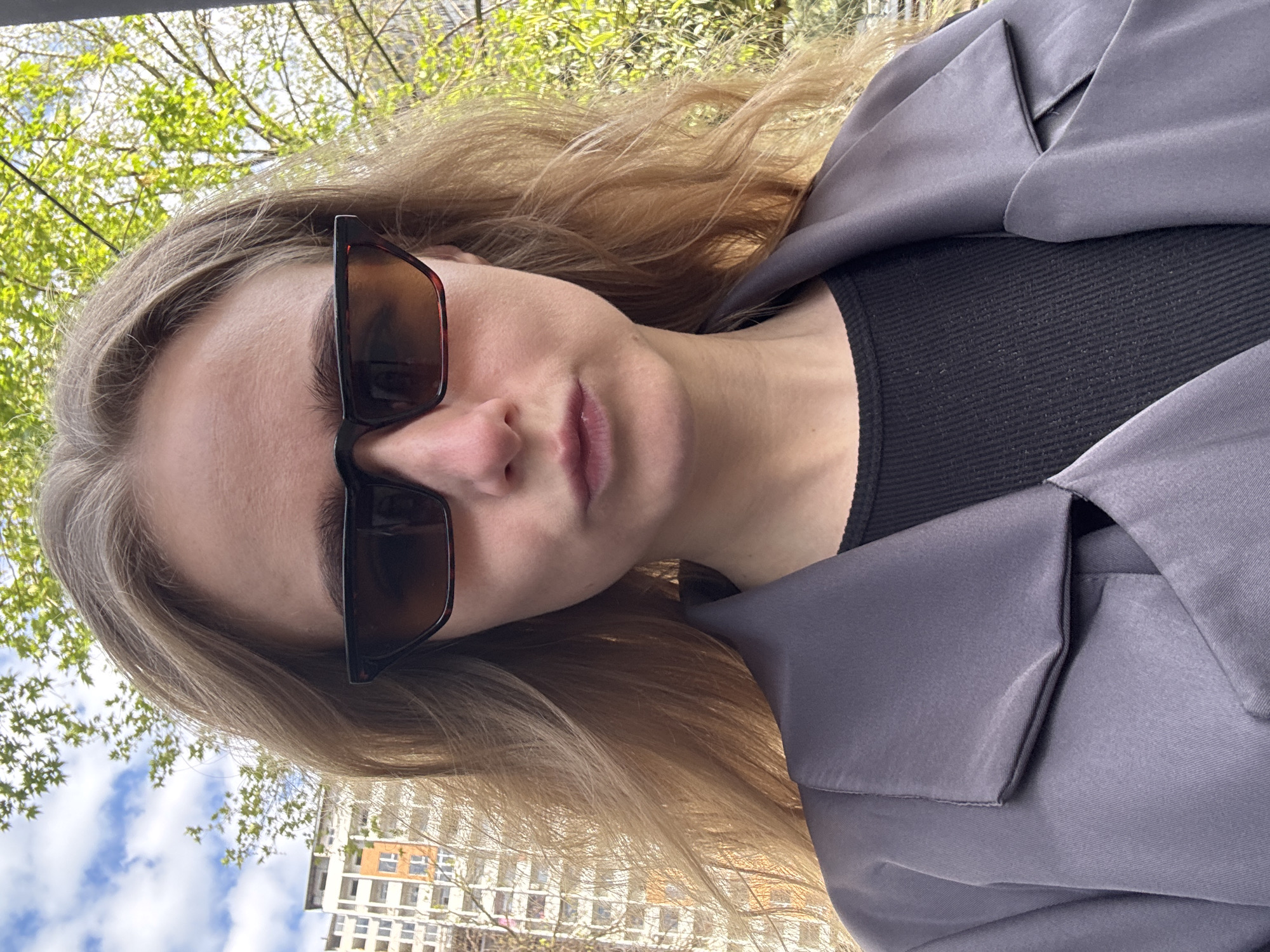 An outdoor portrait, oriented ninety degrees counter-clockwise, features a young woman looking directly at the camera. She has long, wavy blonde or light brown hair that frames her face. Her eyes are obscured by large, rectangular sunglasses with dark brown, possibly tortoise-shell frames and tinted lenses. Her lips are slightly parted, conveying a neutral or serene expression.
She is dressed in a dark grey or muted lavender-grey blazer or jacket with broad lapels, made from a smooth, slightly reflective fabric. Beneath the blazer, a black ribbed top is visible at the neckline.
The background is an outdoor setting, suggesting an urban or suburban environment. To the right side of the image (top in the correctly oriented view), vibrant green trees with fresh spring leaves are visible against a bright, sunlit sky. To the left side (bottom in the correctly oriented view), multiple multi-story buildings with visible windows are set against a partly cloudy blue sky. The bright, natural light indicates it is daytime. No text is visible in the image.