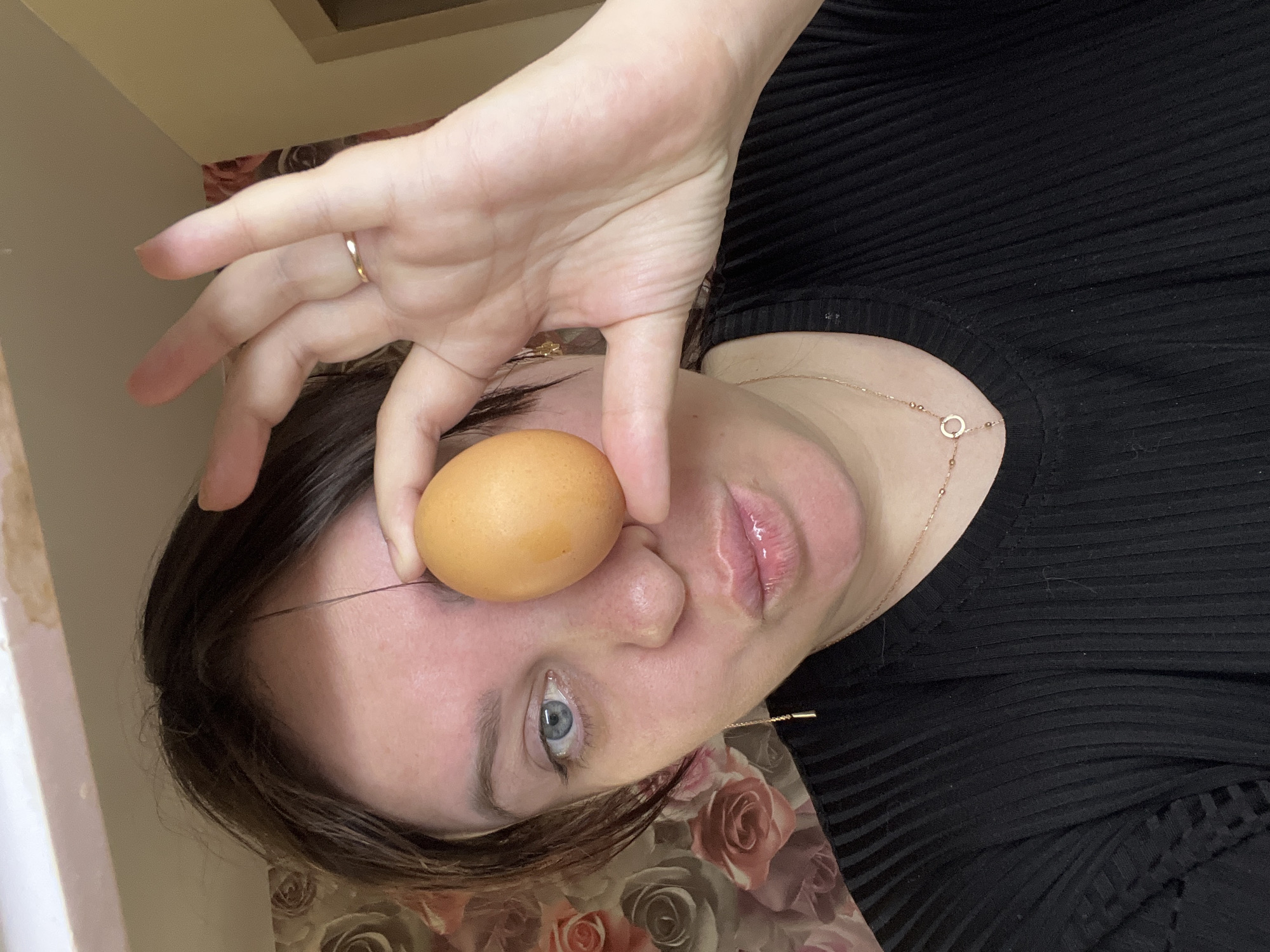 A fair-skinned person with light blue eyes is holding a brown egg in front of their left eye. They are wearing a black, ribbed top and a gold chain necklace. A gold ring is visible on their left hand. The background includes a floral print and a cream-colored wall. There is no information to suggest the location is in Noviy, Russia.