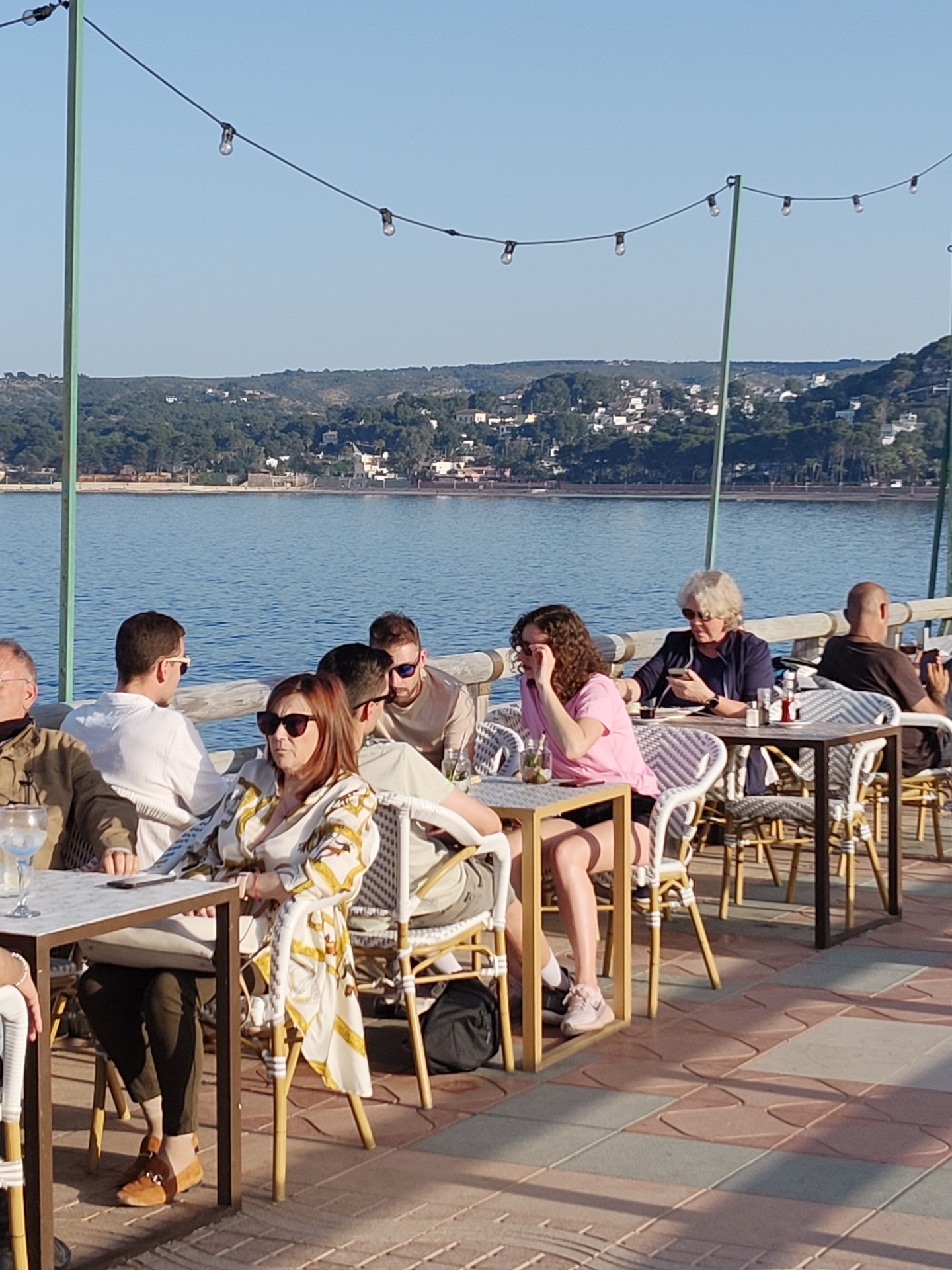 The image shows a group of people dining at an outdoor restaurant in Dénia, Spain, evidenced by the waterfront view. Eight individuals are seated at three tables. The table closest to the camera is occupied by three people. A woman in a patterned dress and sunglasses sits facing the viewer, and two men are seated to her right. The middle table features two women: one in a pink shirt and the other, an older woman, looking at her phone. A man is seated at the third table. A bald man looks toward the water. A string of lights stretches overhead, supported by metal poles. The restaurant is situated on a patterned tile surface, overlooking a body of water with forested hills and white buildings in the background. The sky is clear and blue.